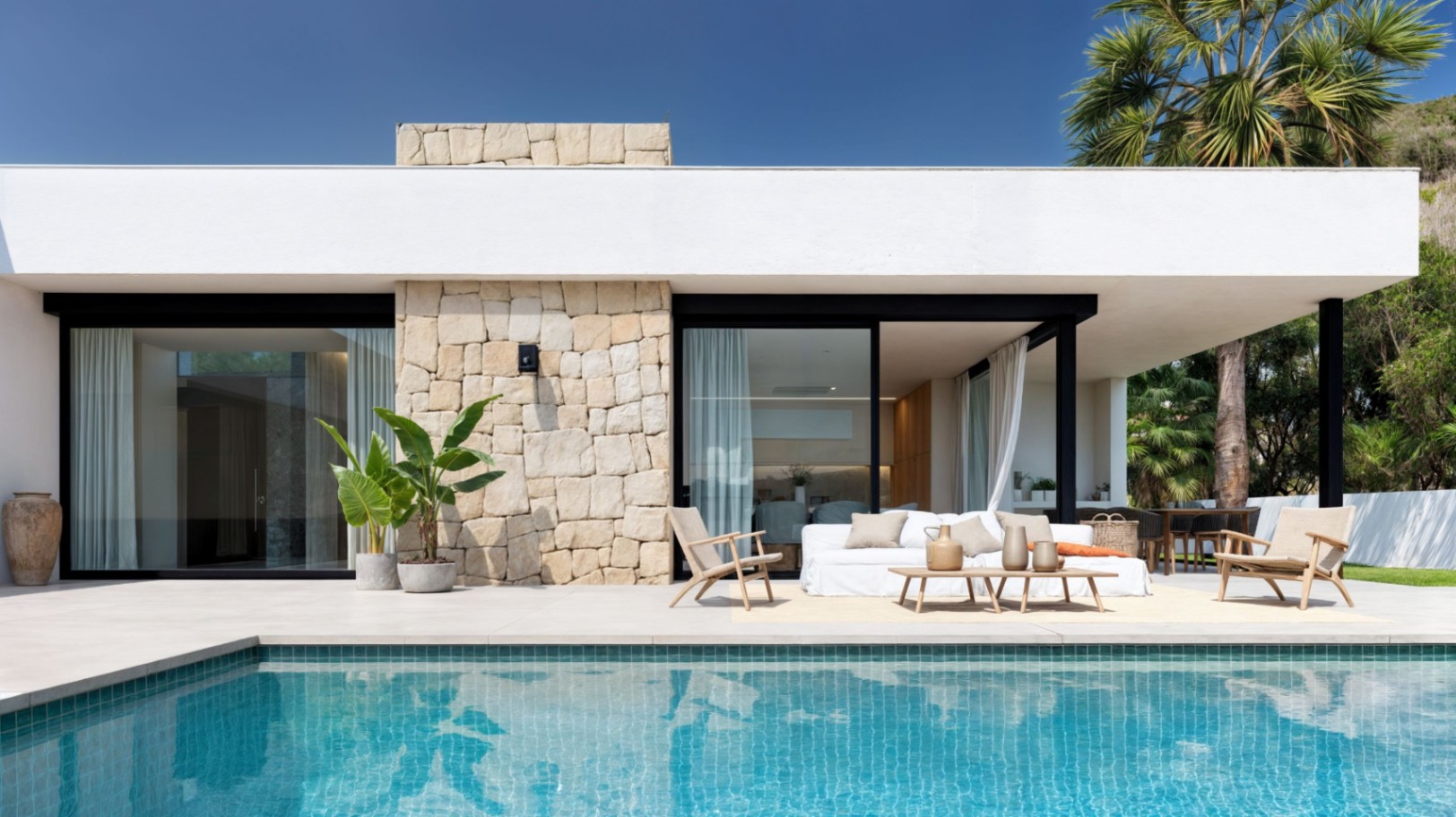 Exterior de villa moderna con piscina de gresite turquesa, muros de piedra natural y zona de estar integrada en una amplia terraza pavimentada.