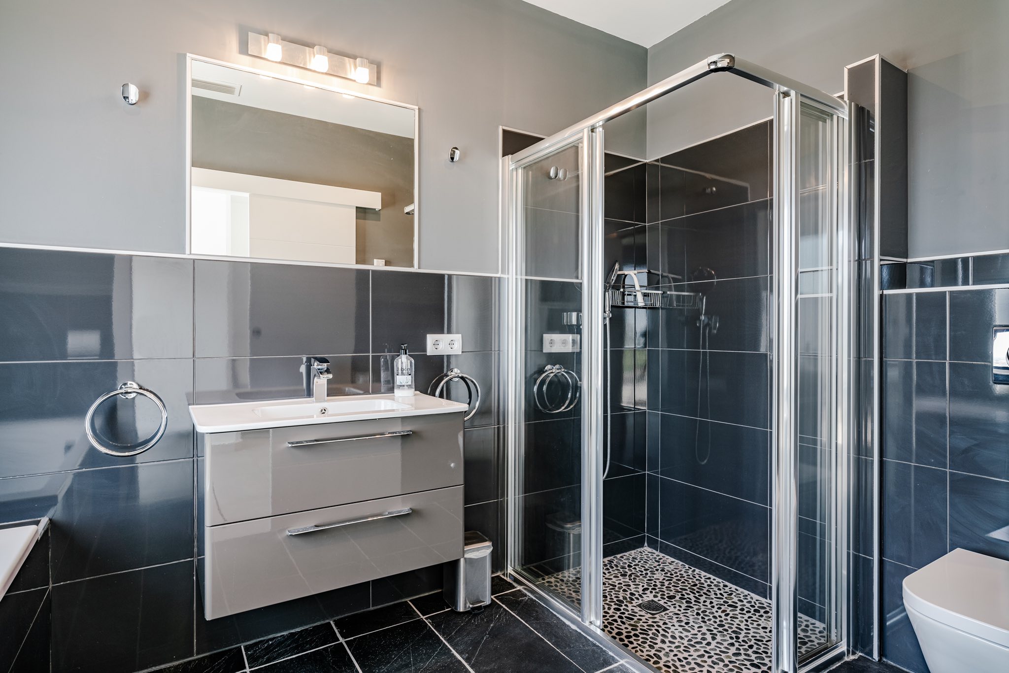 Baño moderno con azulejos gris marengo brillantes, mueble suspendido con lavabo integrado y ducha acristalada con suelo de diseño en guijarros.