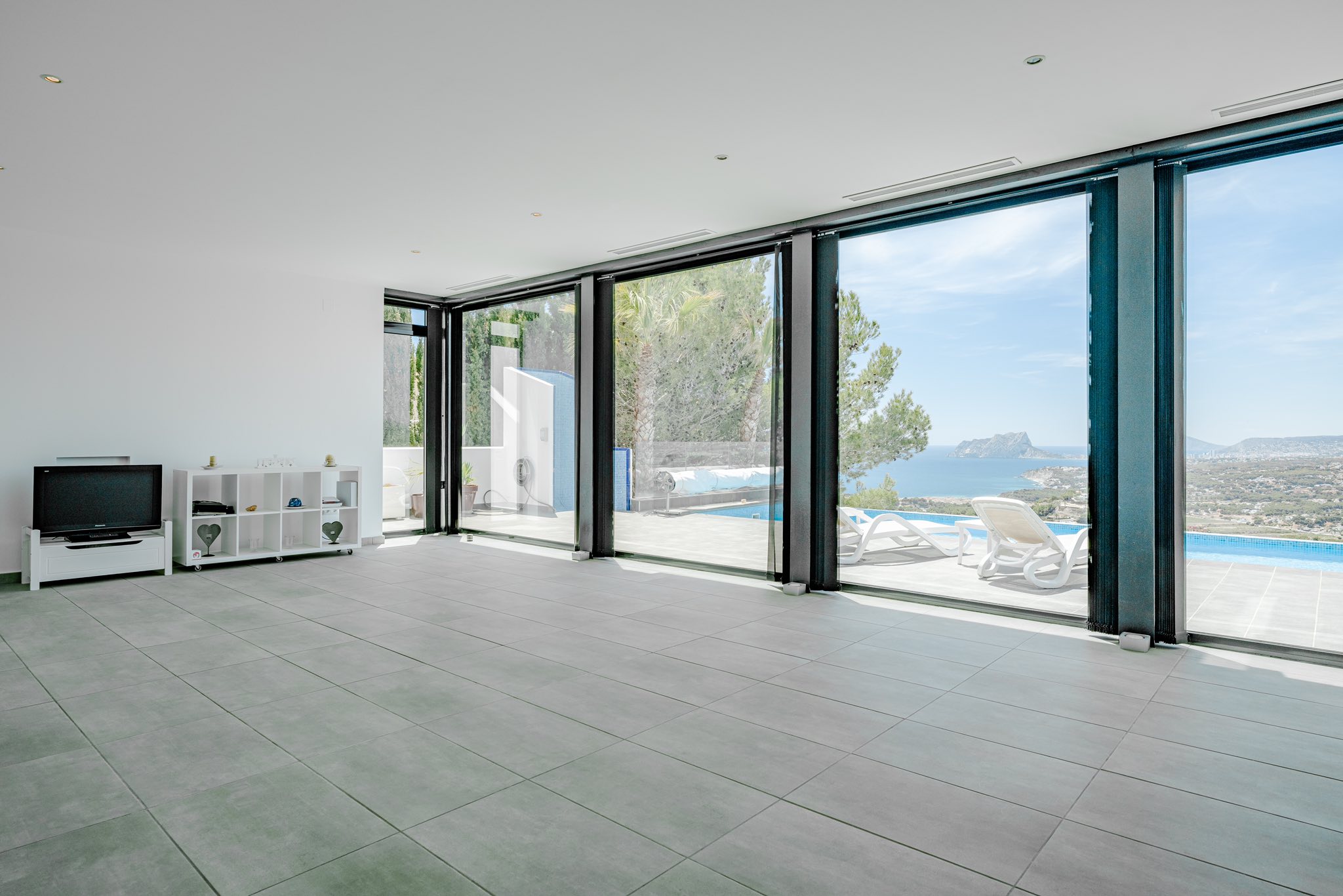 Salón diáfano con suelos de gres gris y amplios ventanales que conectan con una terraza con piscina y vistas panorámicas al mar y al Peñón de Ifach.