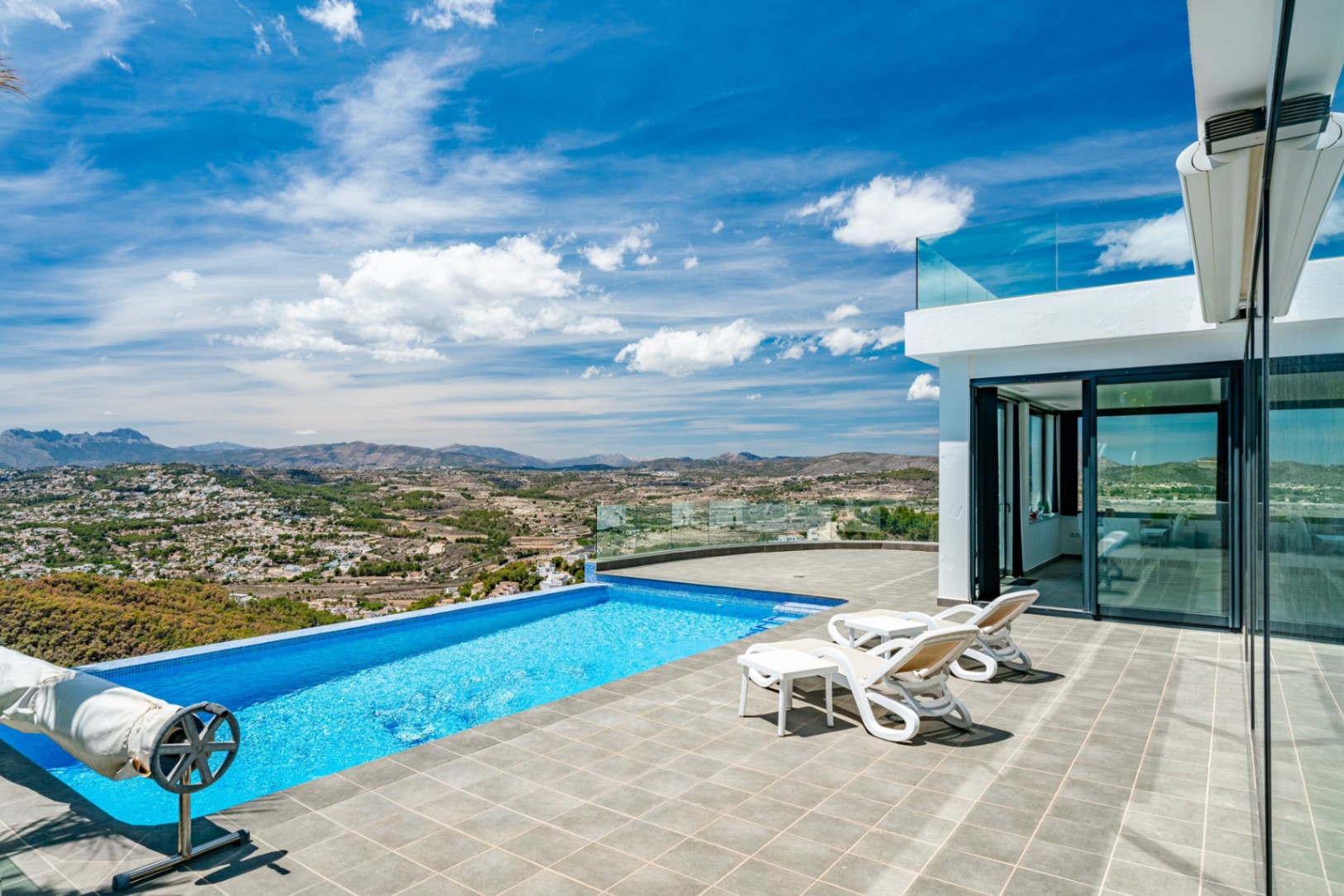 Terraza con piscina infinita y barandillas de cristal, con vistas panorámicas al valle y las montañas. Incluye pavimentos de piedra y tumbonas junto a la villa.