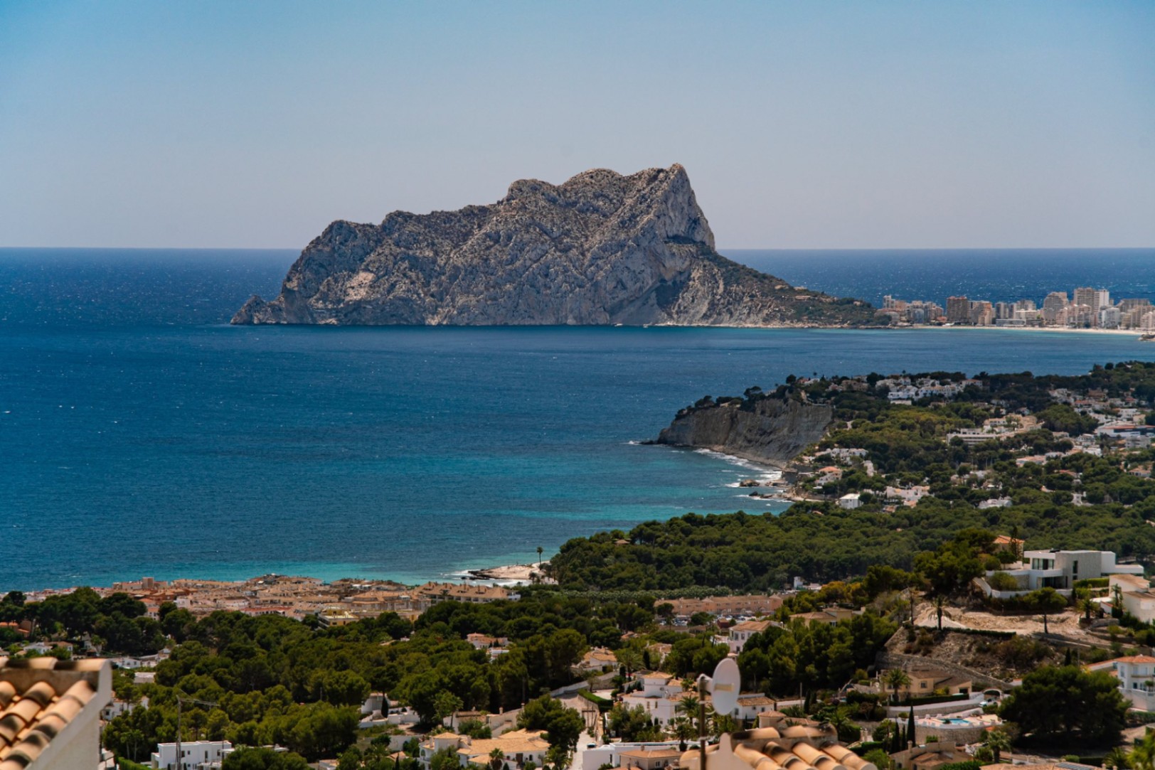 Vista panorámica elevada de la costa mediterránea con el Peñón de Ifach, aguas turquesas y una zona residencial integrada en un frondoso bosque de pinos.