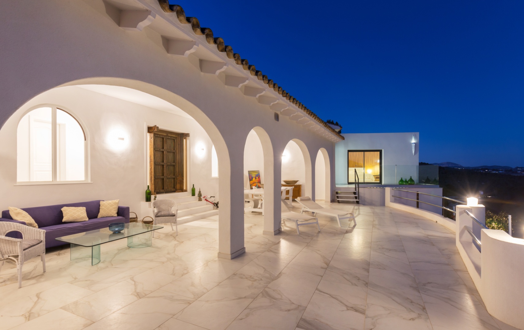 Amplia terraza cubierta al atardecer con arcos blancos, suelo de gres porcelánico veteado, zona de estar con sofá violeta y vistas despejadas.
