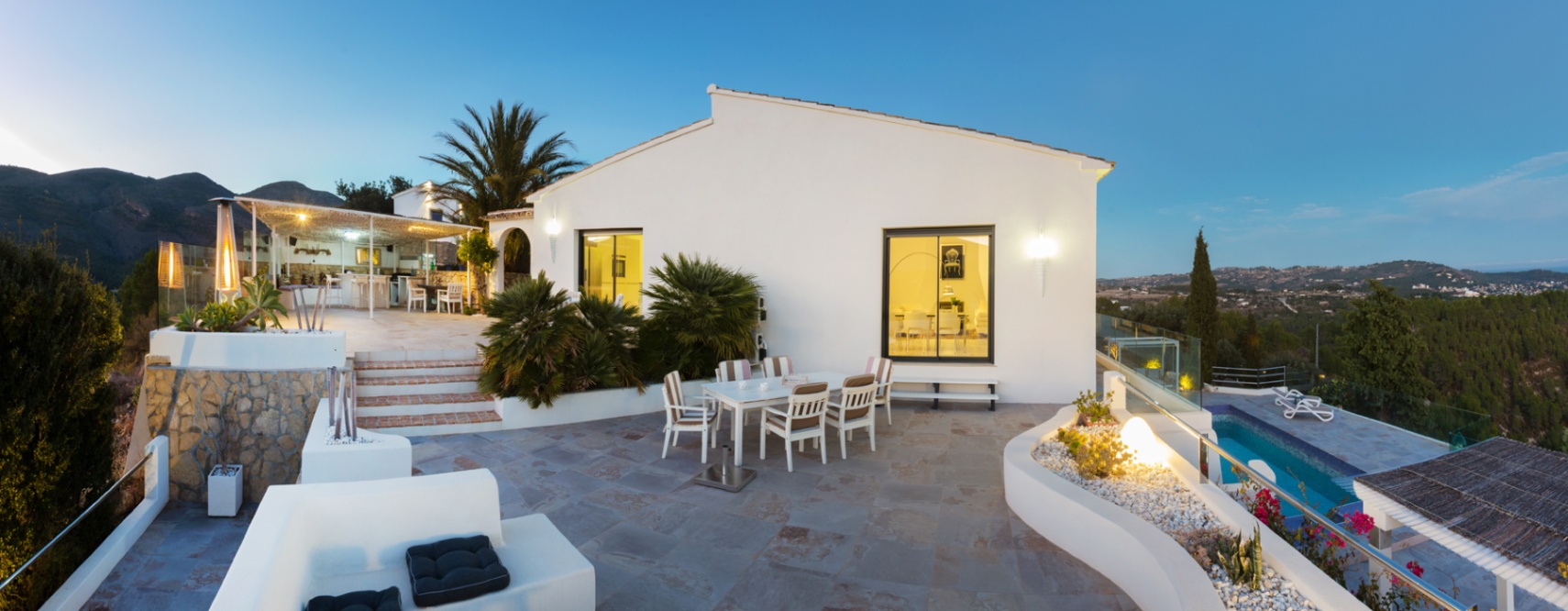 Amplia terraza de piedra al atardecer con zona de comedor, jardineras integradas y vistas panorámicas al valle junto a una villa blanca de estilo mediterráneo.