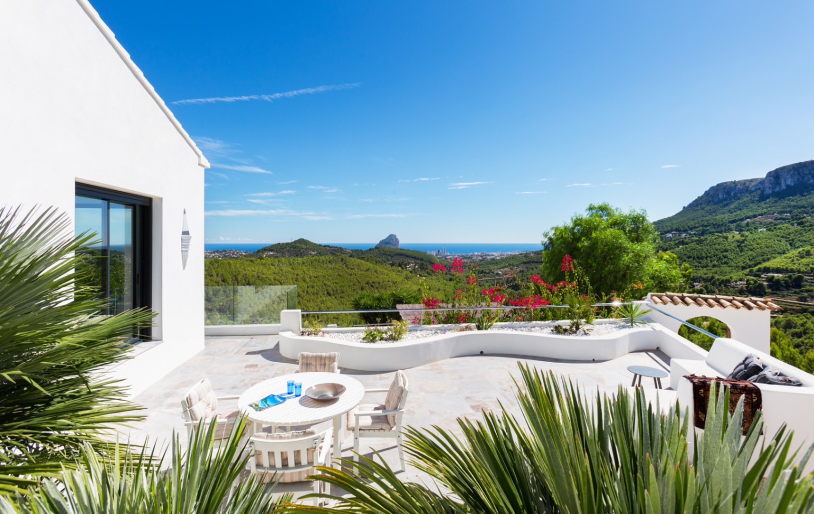 Amplia terraza de piedra con comedor exterior y jardineras curvas, que ofrece vistas panorámicas de la costa mediterránea y el Peñón de Ifach.