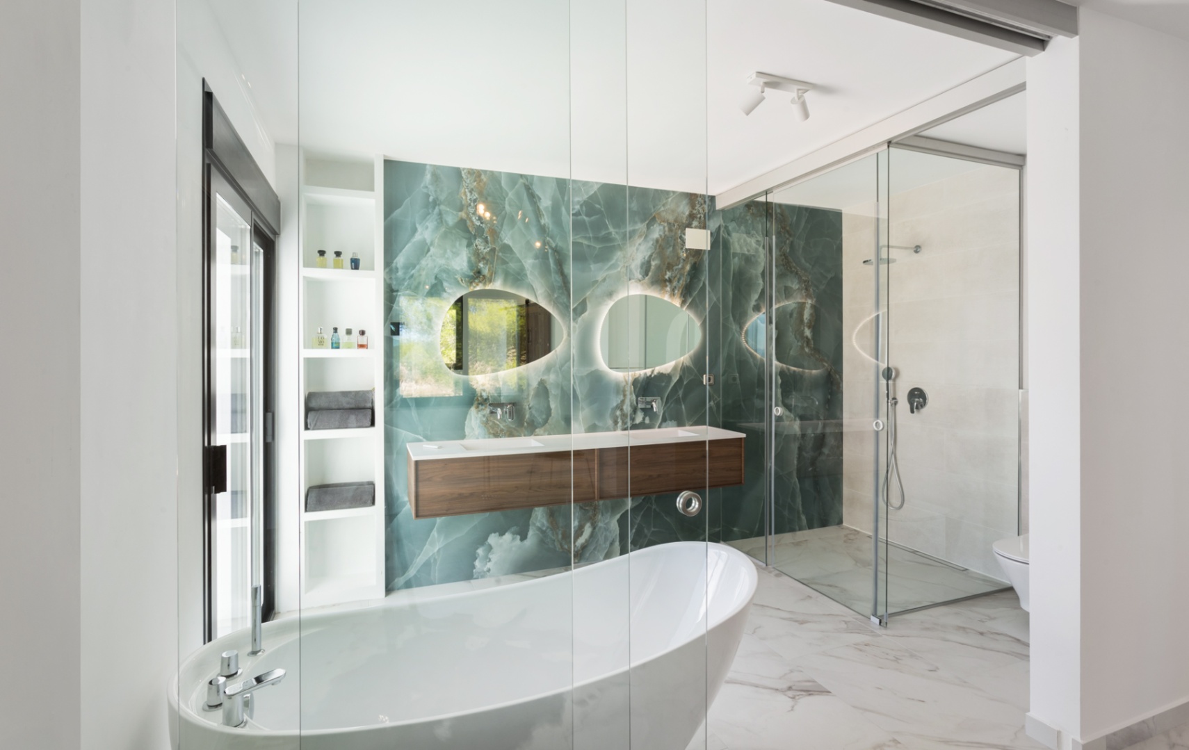 Baño de diseño con pared frontal de ónix verde, bañera exenta blanca y mueble de lavabo doble en madera oscura. Incluye espejos orgánicos retroiluminados y ducha de cristal.