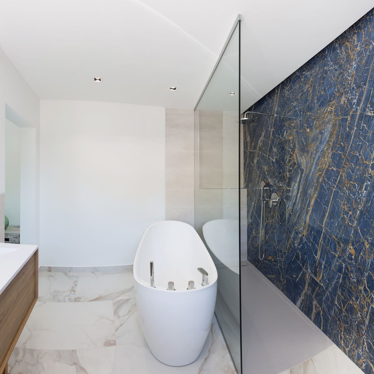 Baño moderno con bañera exenta ovalada, ducha a ras de suelo con pared de mármol azul veteado y suelos de mármol blanco con detalles dorados.