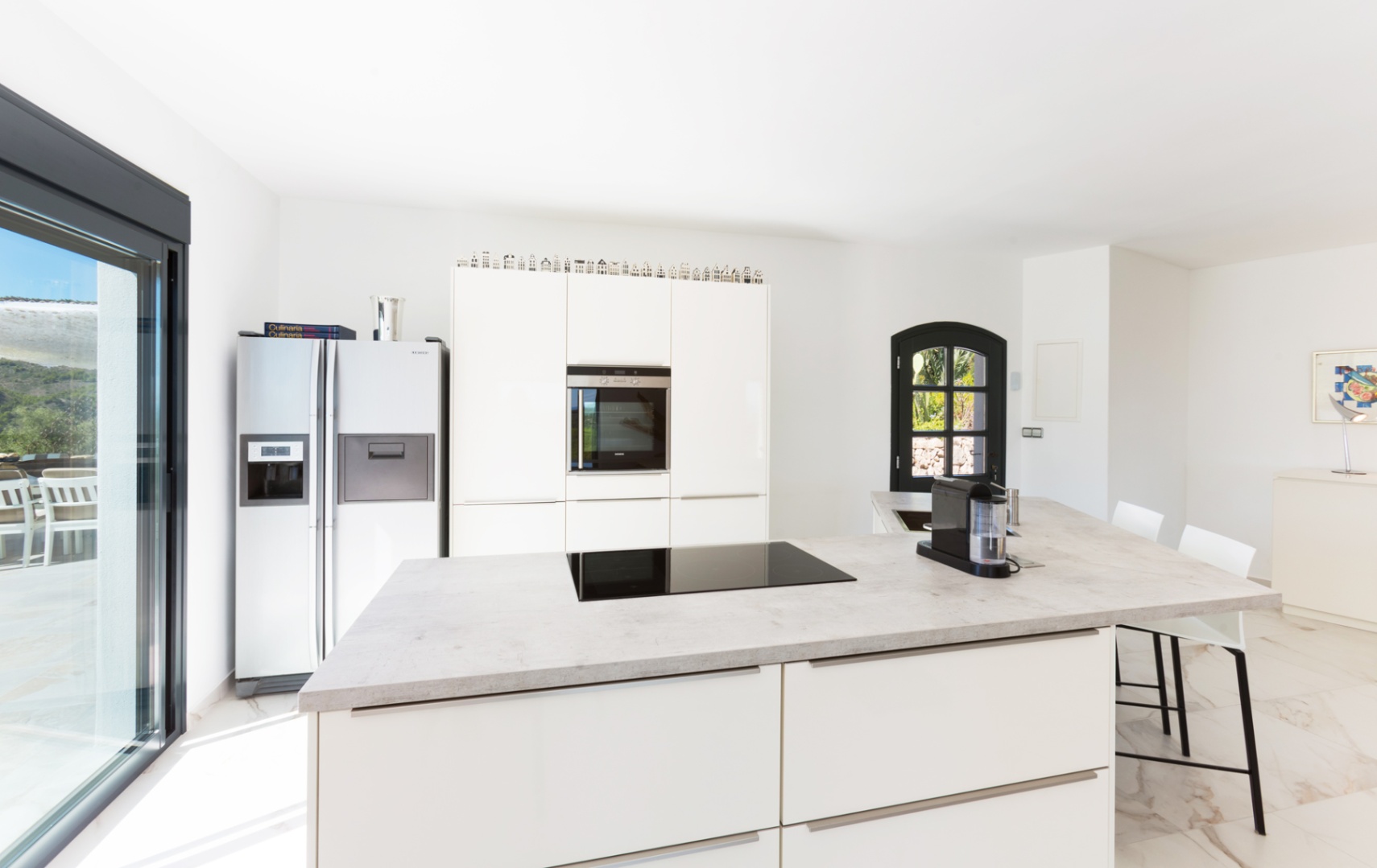 Cocina moderna con isla central de efecto piedra, placa de inducción, armarios blancos y frigorífico americano. Suelo de mármol y acceso directo al exterior.