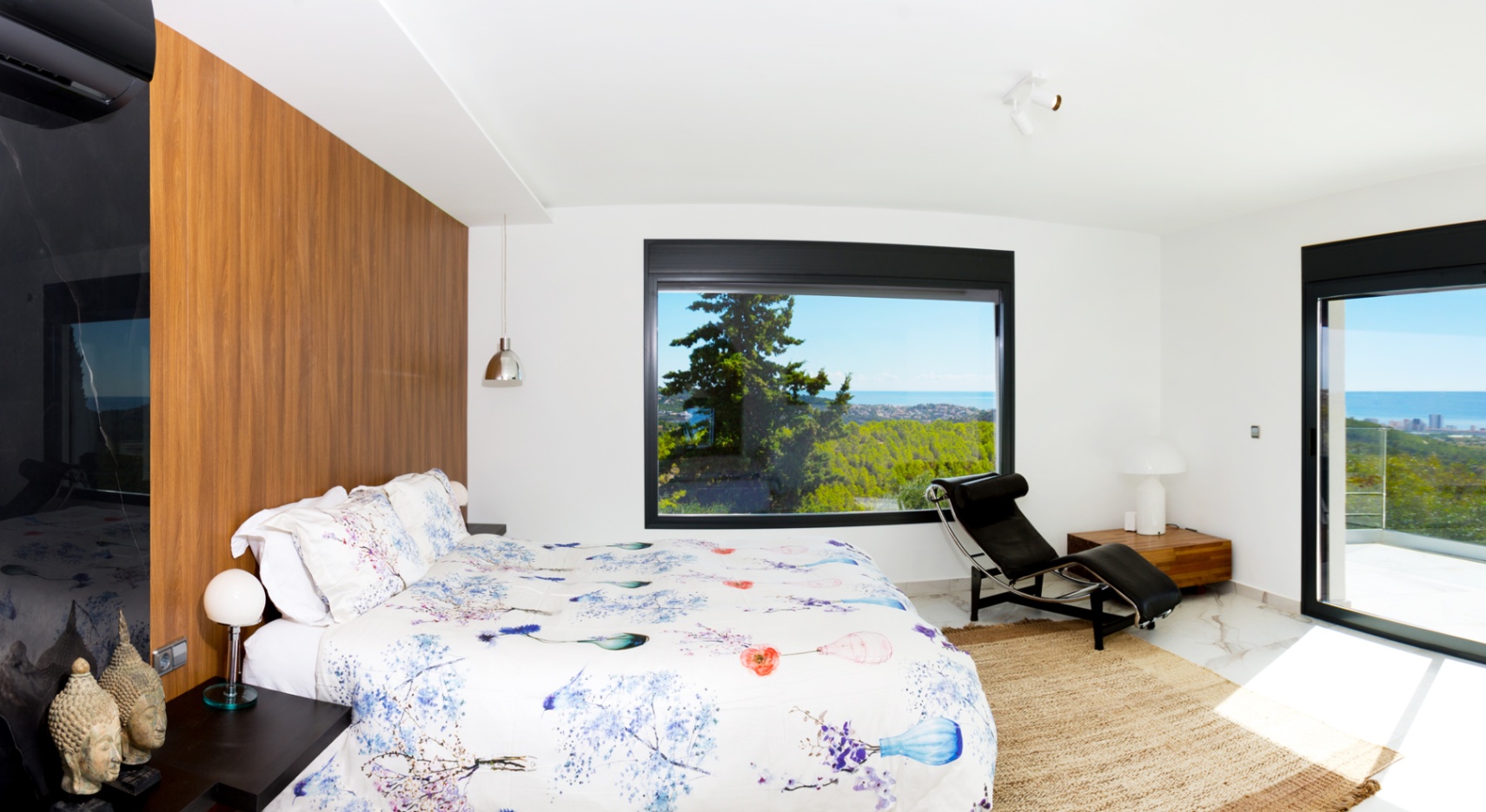 Dormitorio moderno con pared de acento en madera, suelos de mármol y un gran ventanal con vistas panorámicas al mar y al bosque. Incluye una chaise longue de diseño.