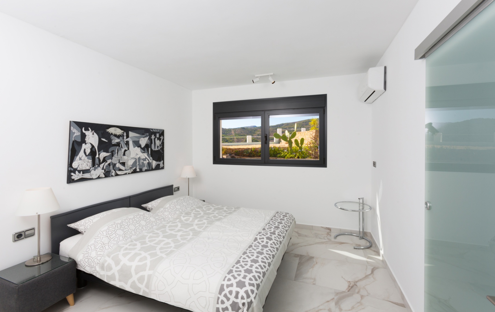 Dormitorio moderno con suelo porcelánico efecto mármol, cuadro monocromático y ventana de marco negro con vistas a una terraza con cactus y montañas.