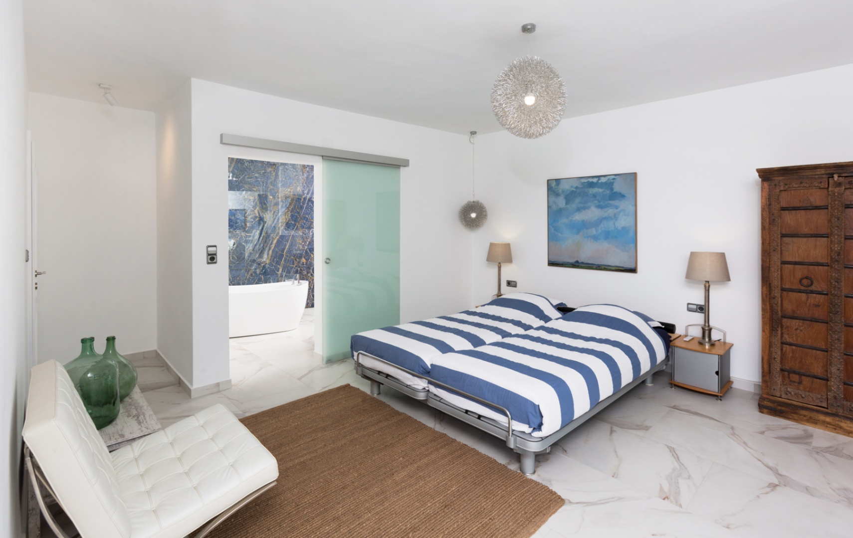 Dormitorio moderno con suelos de mármol, cama con textiles a rayas y puerta corredera de cristal hacia un baño en suite con bañera exenta y pared de piedra azul veteada.