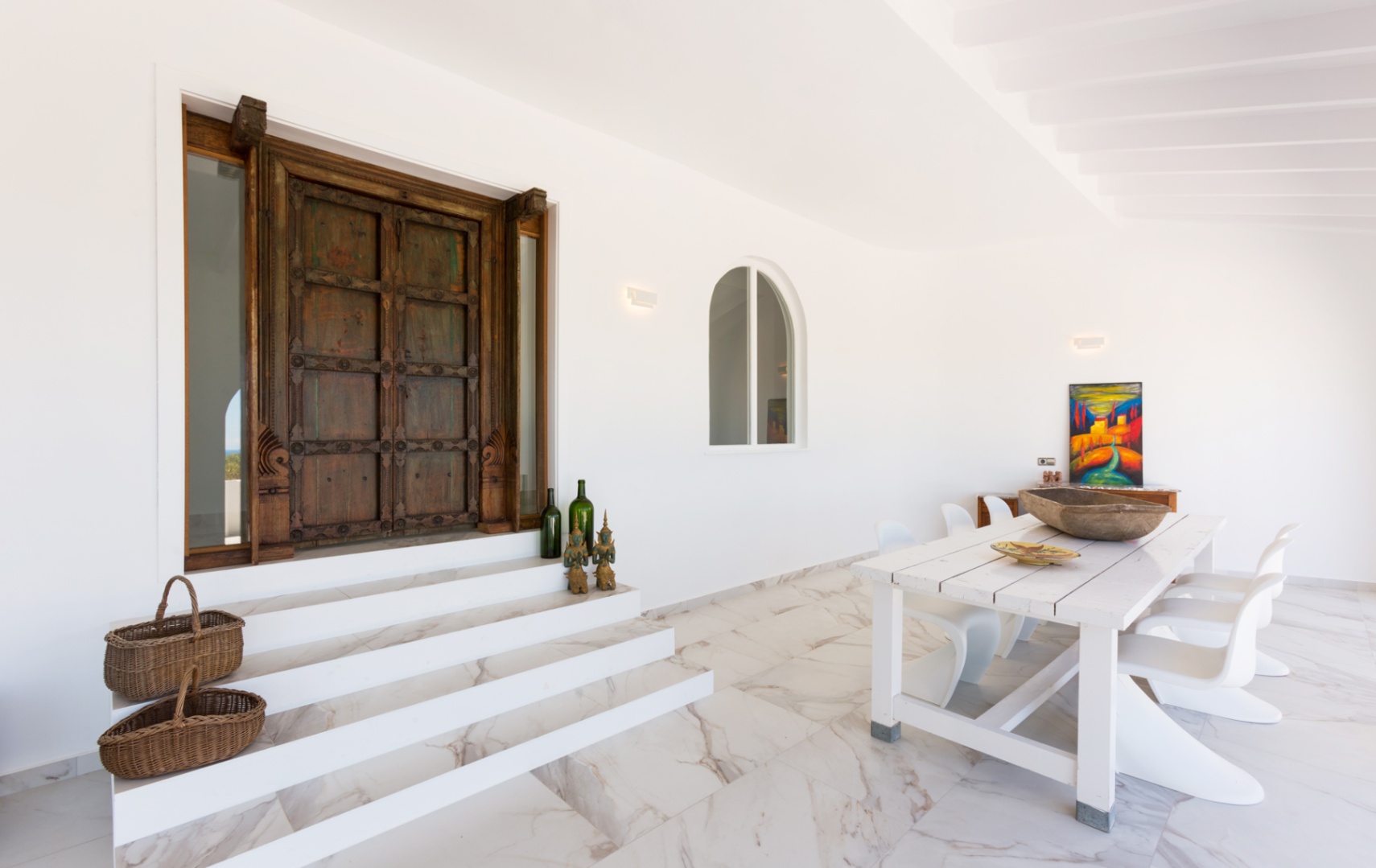 Entrada con puerta de madera tallada antigua, escalones de mármol blanco y zona de comedor exterior con mesa rústica y sillas de diseño contemporáneo.