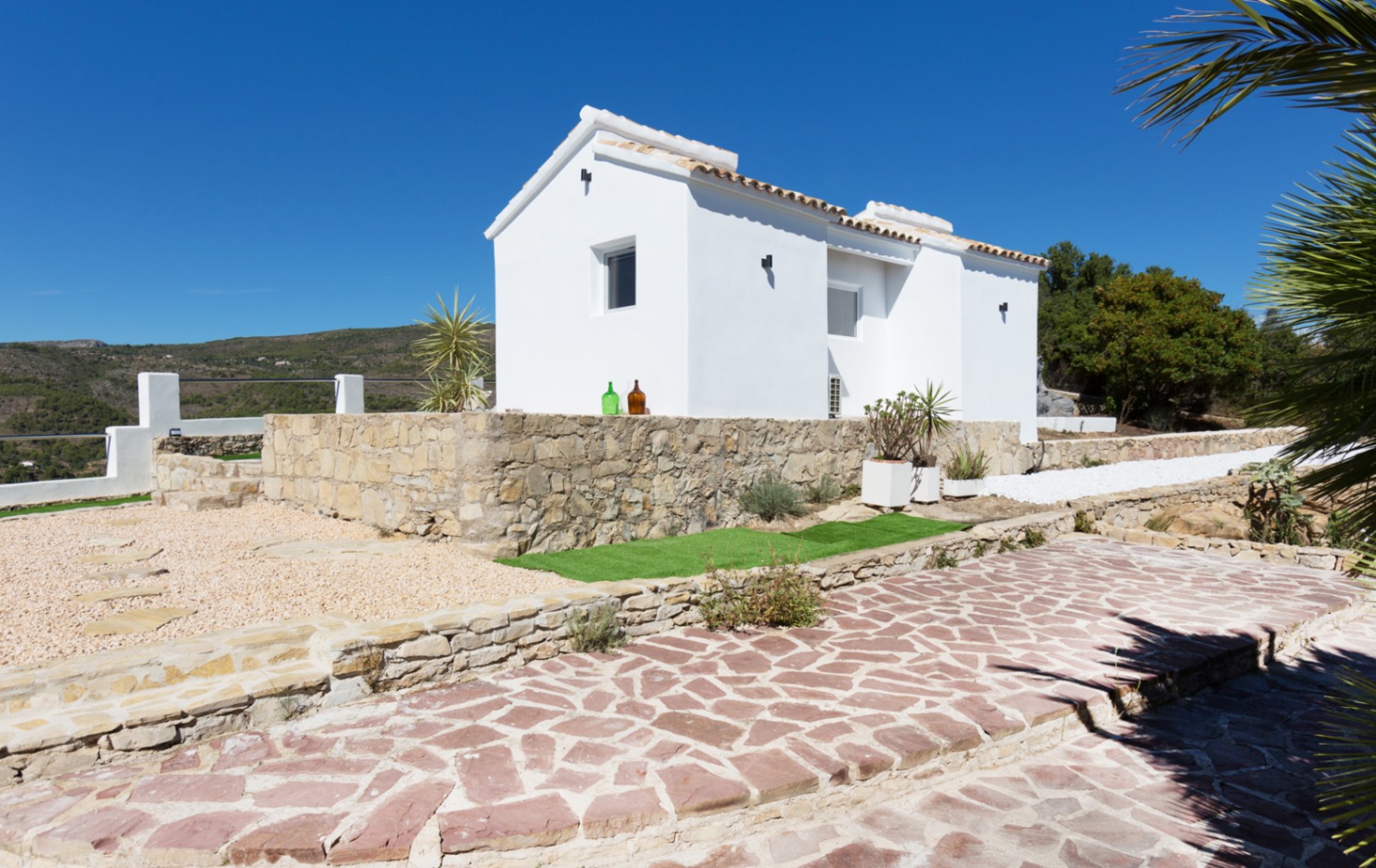 Exterior de una villa blanca con muros de piedra natural, tejado de teja cerámica y un camino pavimentado rodeado de zonas de grava y vegetación mediterránea.