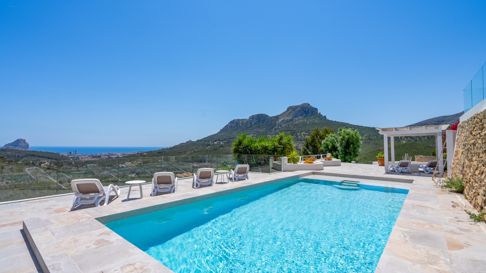 Piscina rectangular con terraza de piedra natural, barandillas de cristal y vistas panorámicas a la costa mediterránea y las montañas circundantes.
