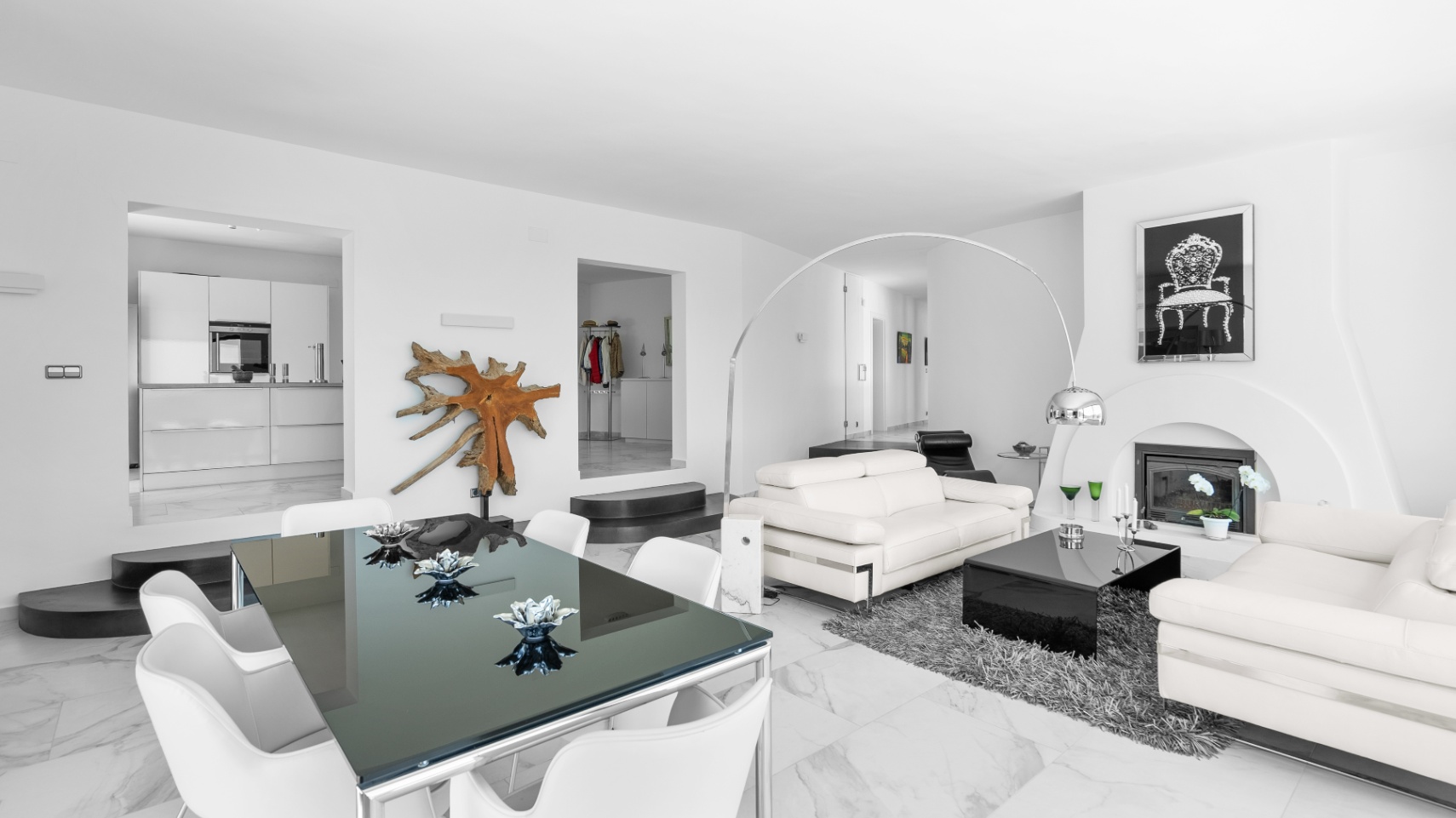 Salón comedor diáfano con suelos de mármol, sofás de cuero blanco, chimenea de diseño y mesa de cristal, con acceso visual a la cocina minimalista.
