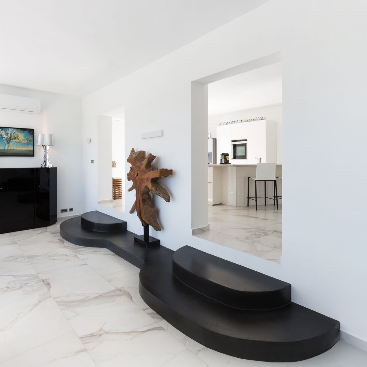 Salón moderno con suelos de mármol blanco y una plataforma curva en negro. Una escultura de raíz natural decora el acceso a la cocina minimalista de tonos blancos.
