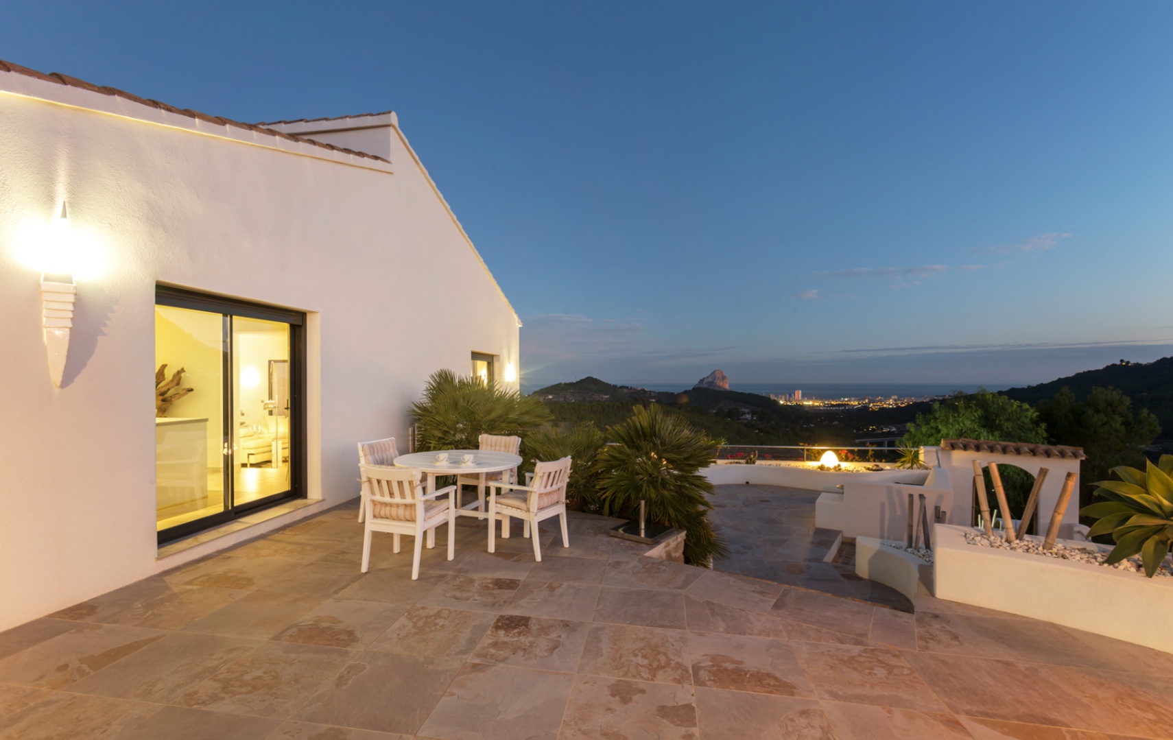 Terraza con suelo de piedra natural, mobiliario de comedor blanco y vistas panorámicas de la costa de Calpe y el Peñón de Ifach al anochecer.