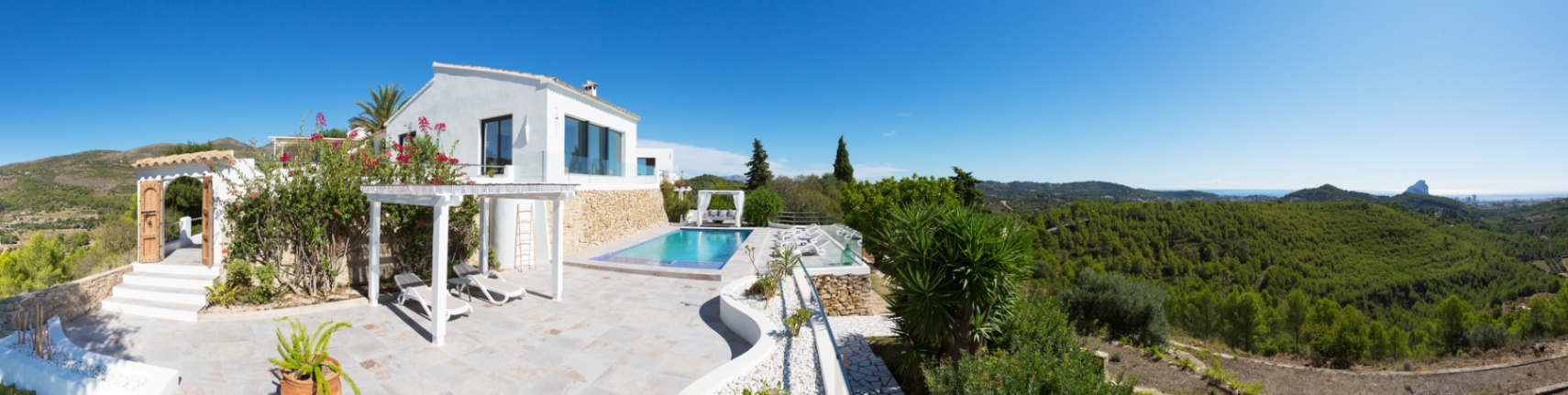Terraza panorámica con piscina, pérgola blanca y suelos de piedra, ofreciendo vistas despejadas al valle y al mar Mediterráneo con el Peñón de Ifach al fondo.
