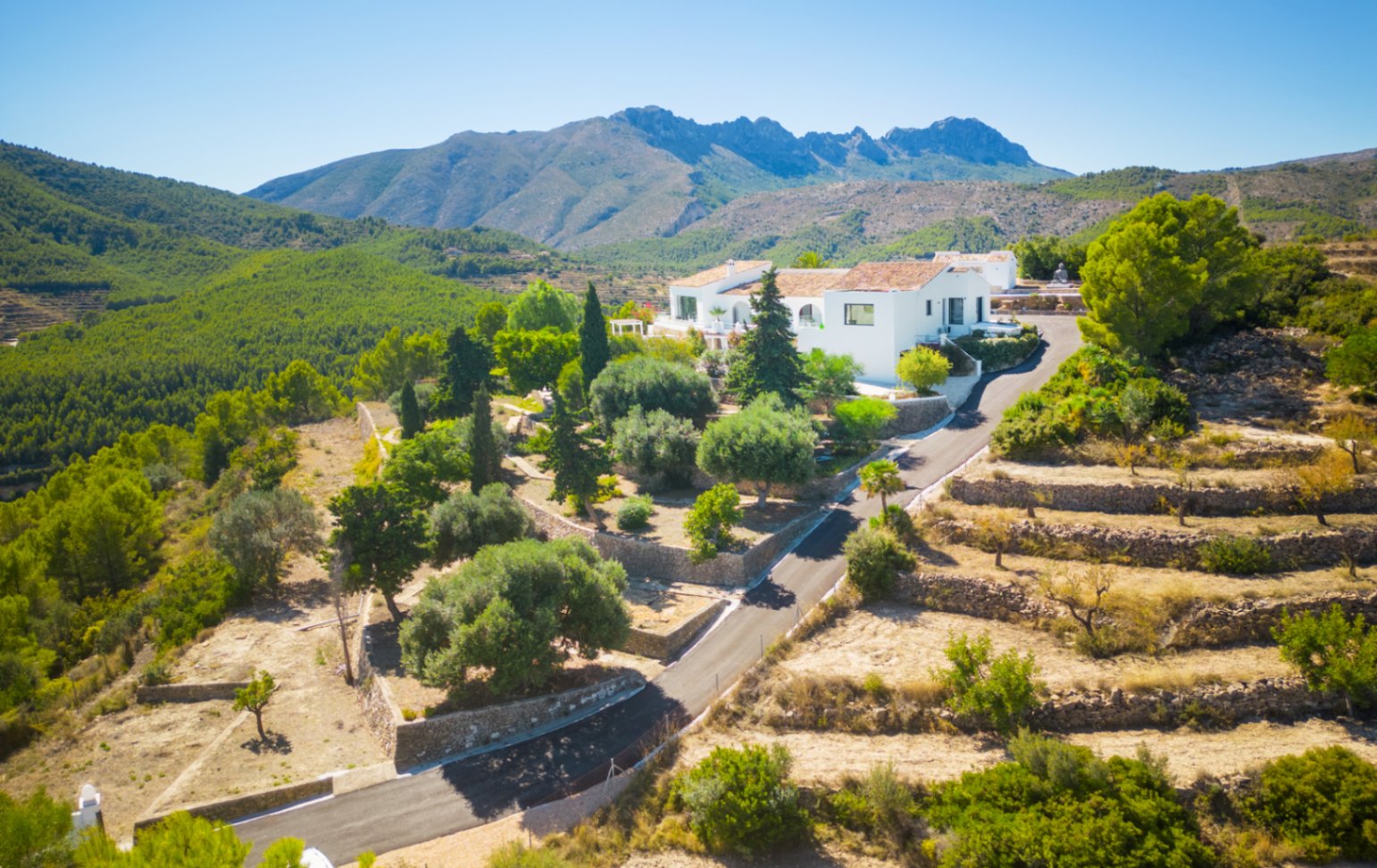 Vista aérea de una villa blanca situada en un entorno montañoso con bancales de piedra, jardines mediterráneos y vistas panorámicas al bosque de pinos.