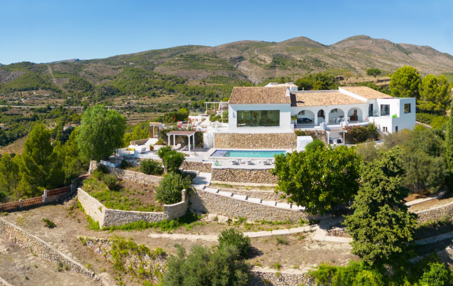 Vista elevada de una villa blanca mediterránea con jardines aterrazados de piedra, piscina privada y porche de arcos frente a un entorno montañoso natural.