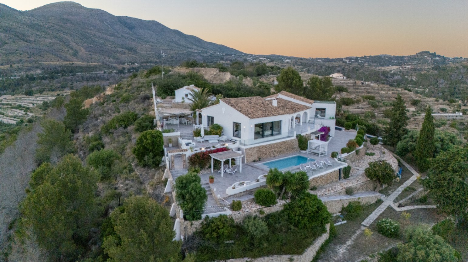 Vista elevada de una villa mediterránea blanca con muros de piedra aterrazados, piscina y varias zonas de descanso exteriores frente a un paisaje montañoso.