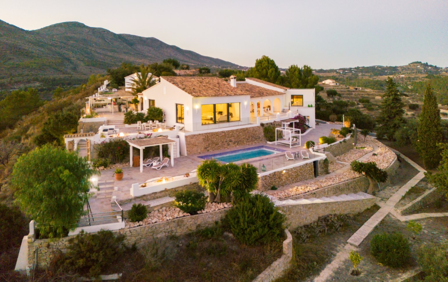 Vista elevada de una villa mediterránea con fachadas blancas, tejados de terracota y terrazas en varios niveles con piscina y muros de piedra natural.