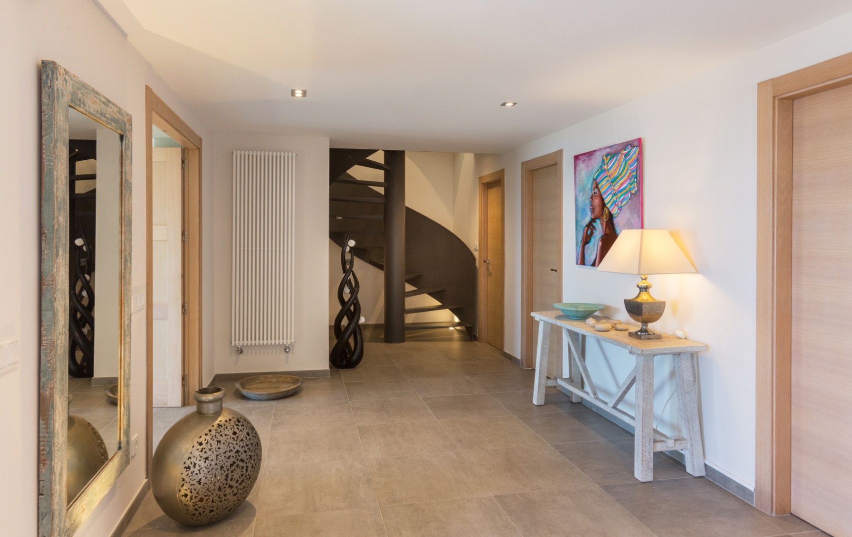 Amplio recibidor con suelos de gres gris, escalera de caracol metálica, puertas de madera clara y una mesa consola rústica con lámpara y arte decorativo.