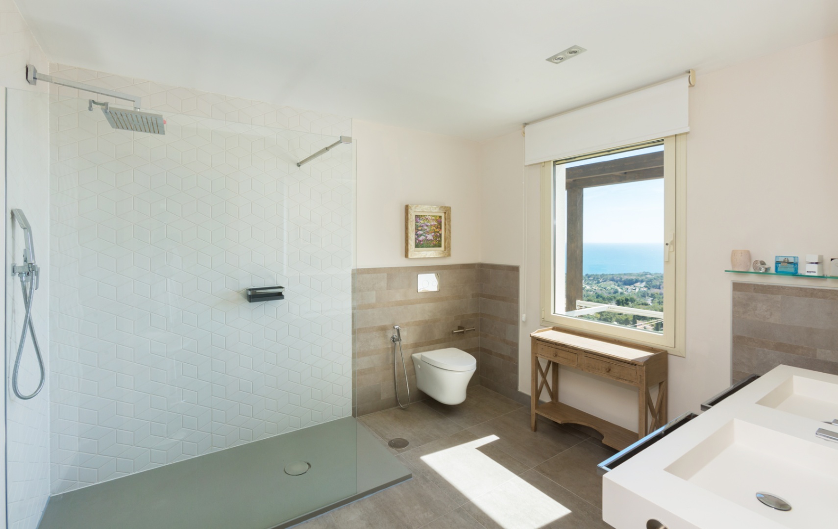 Baño moderno con ducha a ras de suelo y azulejos hexagonales, lavabo doble y un gran ventanal con vistas panorámicas al mar y la costa.