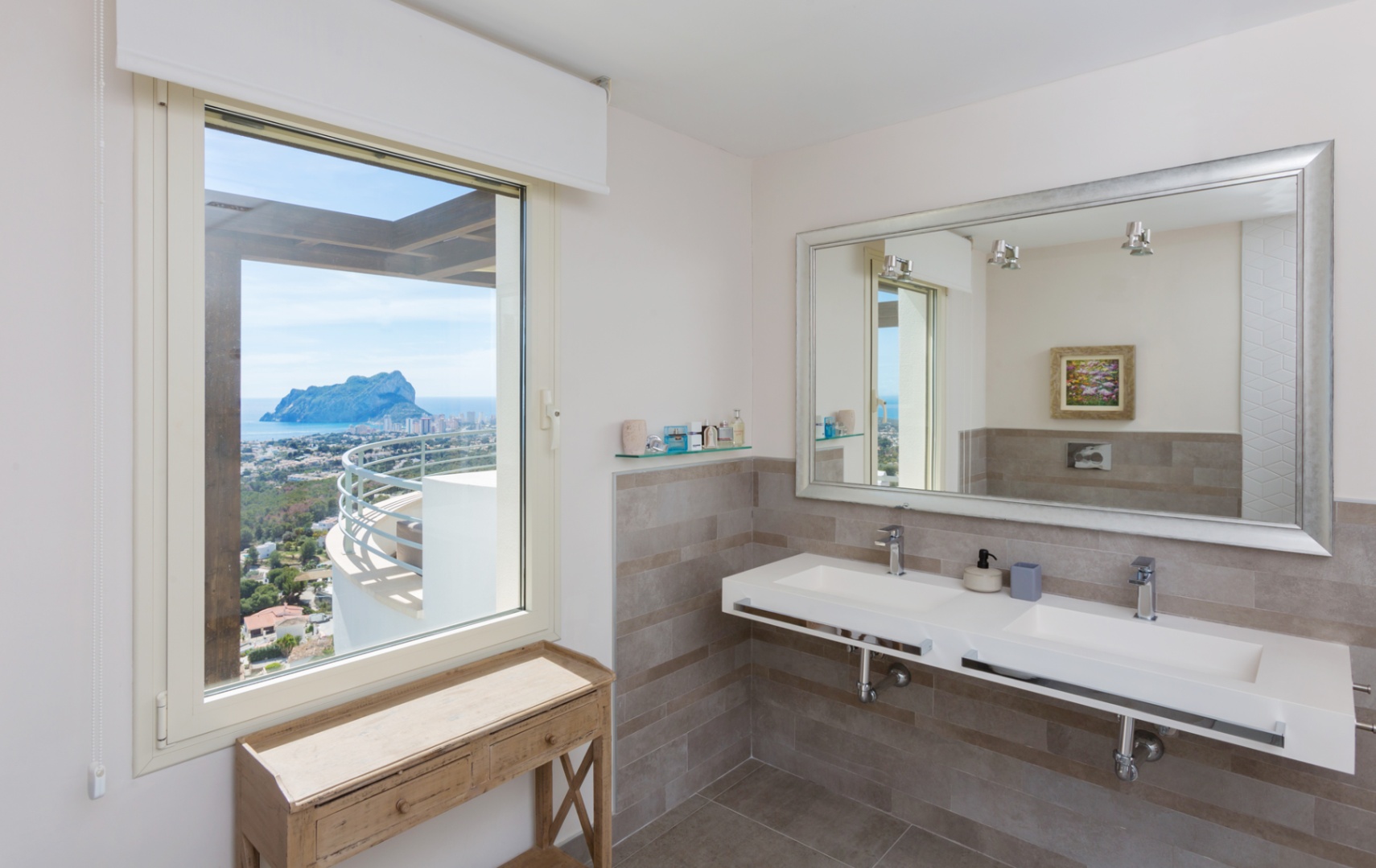 Baño moderno con lavabo doble suspendido, espejo de marco plateado y ventana con vistas panorámicas al Peñón de Ifach y el mar Mediterráneo.