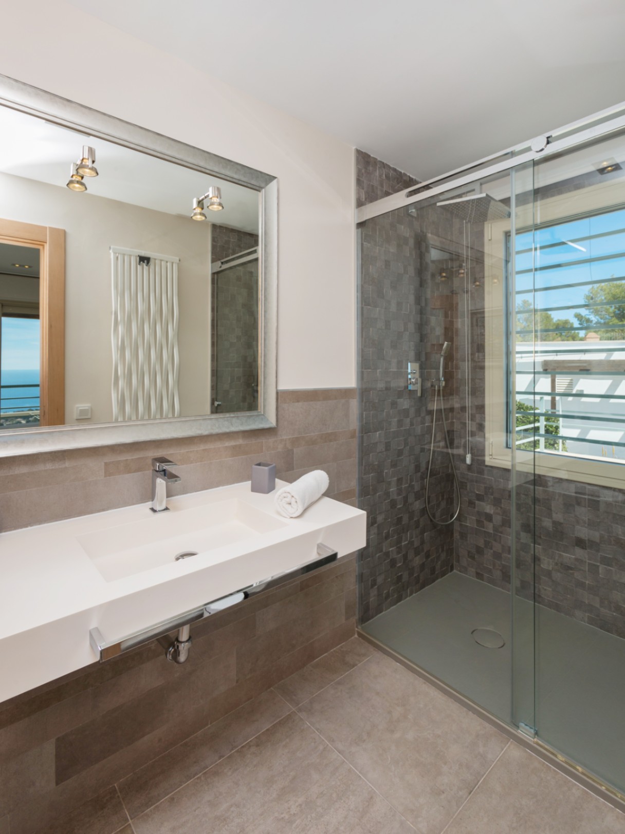 Baño moderno con lavabo integrado blanco, gran espejo de marco plateado y ducha de obra con azulejos de mosaico gris y luz natural.