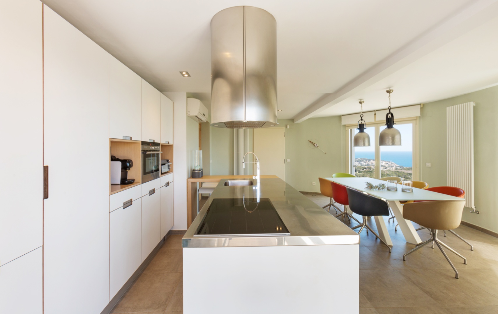Cocina moderna de concepto abierto con isla blanca, encimera de acero inoxidable, campana cilíndrica y comedor con vistas al mar a través de ventanales.