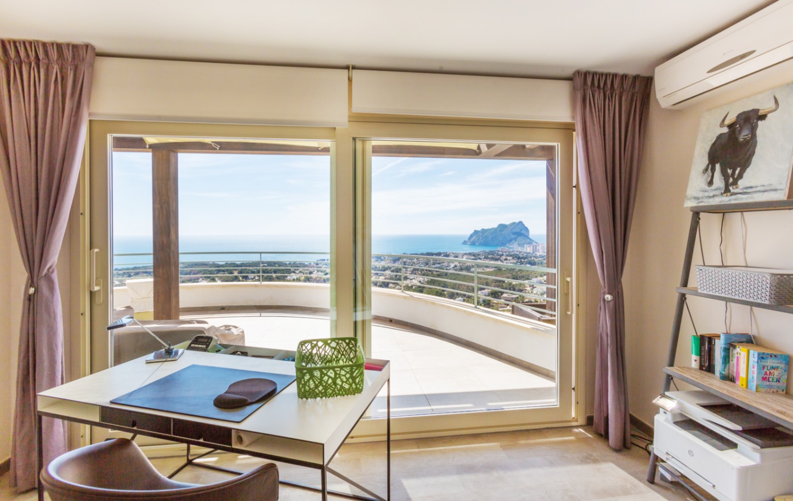 Despacho con escritorio moderno y estanterías, equipado con grandes ventanales que dan acceso a una terraza con vistas panorámicas al mar y al Peñón de Ifach.