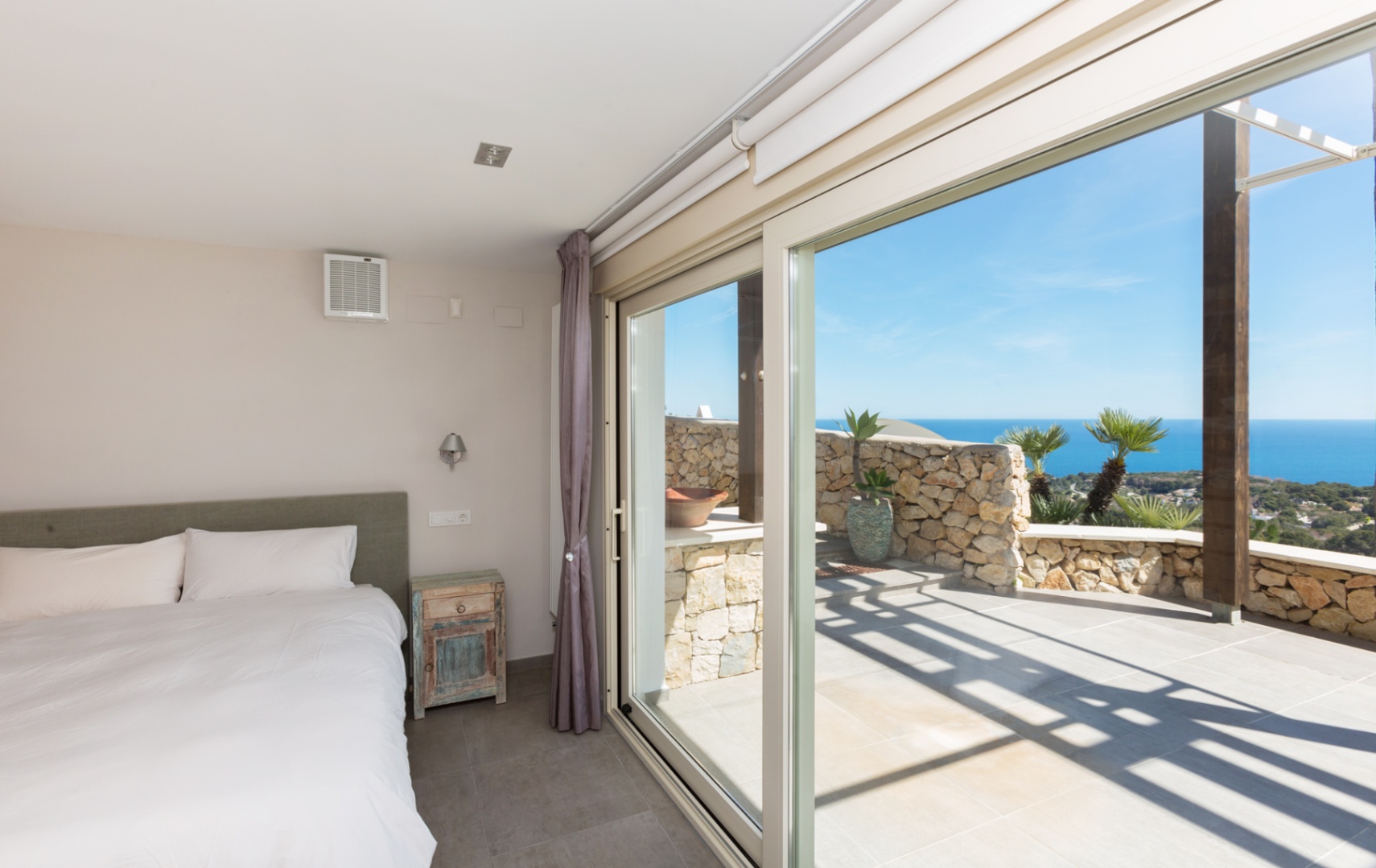 Dormitorio con amplios ventanales correderos que conectan con una terraza de piedra y ofrecen vistas panorámicas al mar Mediterráneo.