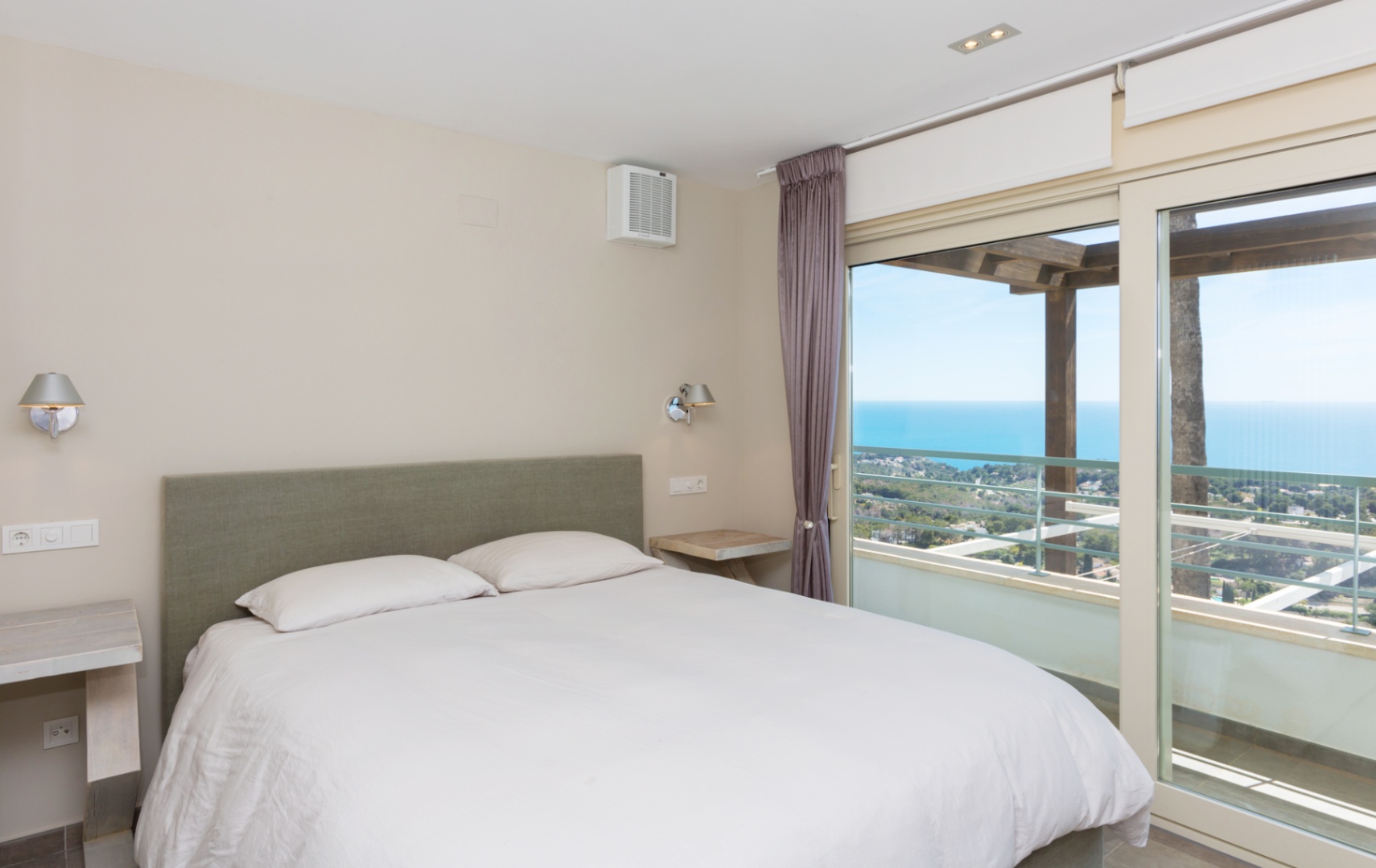 Dormitorio con cabecero tapizado en verde y grandes ventanales correderos que dan acceso a una terraza con pérgola de madera y vistas panorámicas al mar.