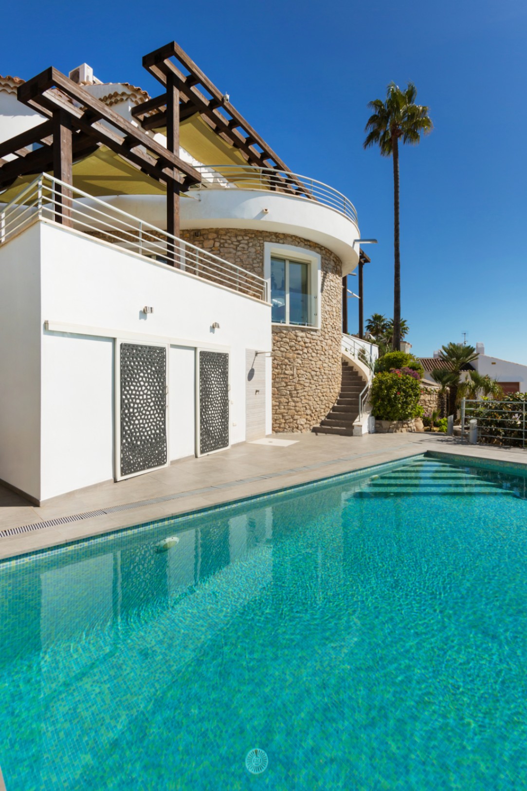 Exterior con piscina de azulejos turquesa junto a una villa de fachada blanca, muro circular de piedra natural, pérgola de madera y paneles metálicos decorativos.
