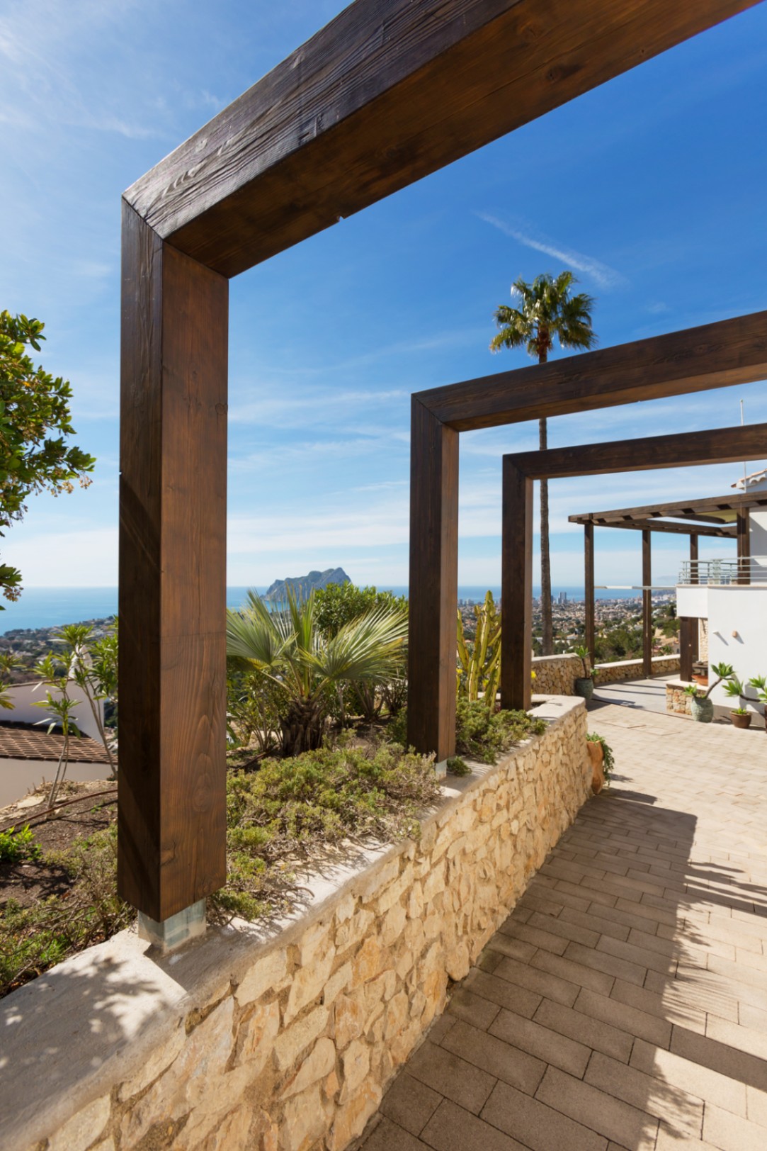 Pasarela exterior con pérgolas de madera oscura, muro de piedra natural y vistas panorámicas al Mediterráneo con el Peñón de Ifach.