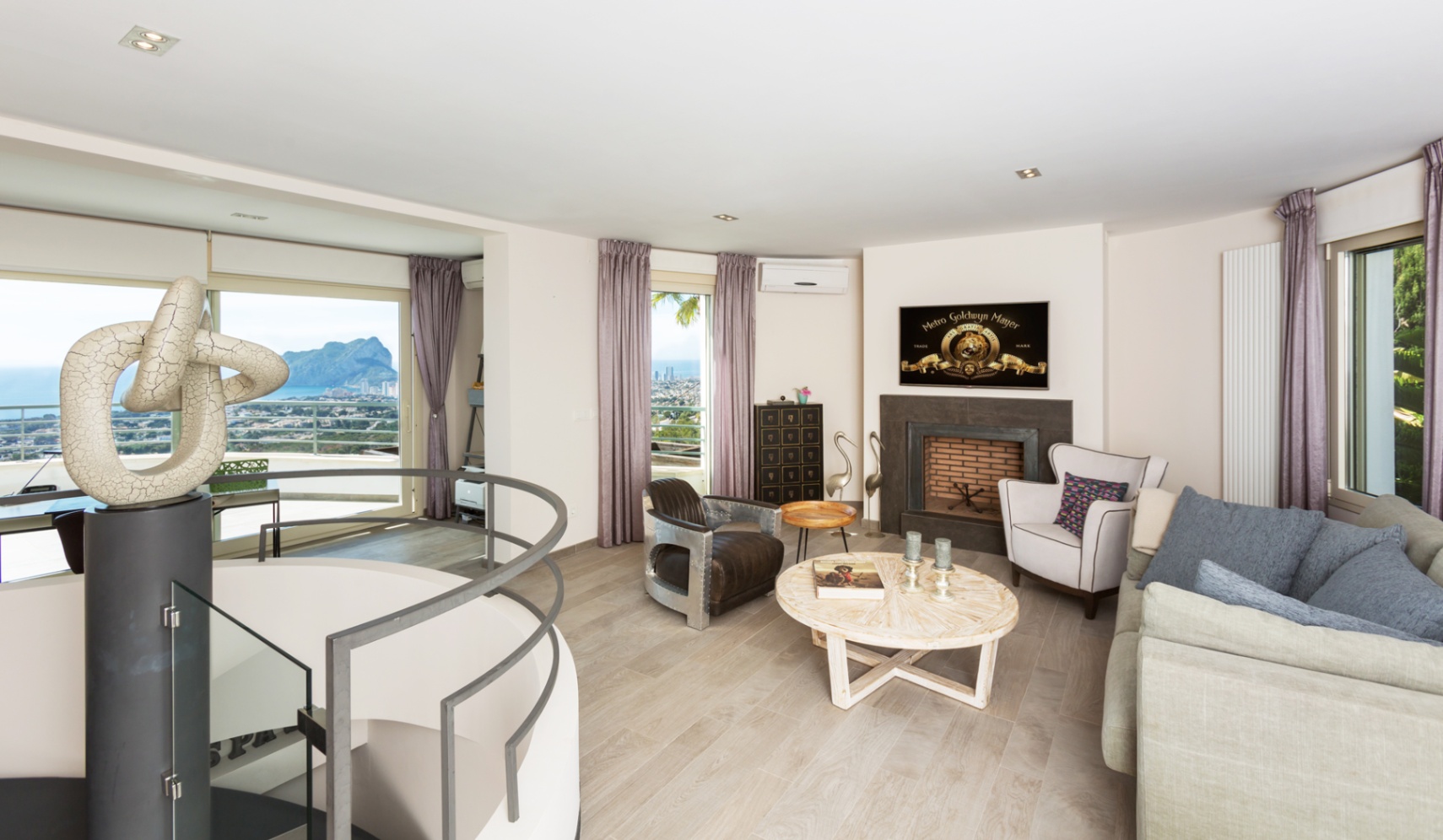 Salón contemporáneo con chimenea, escalera de caracol con escultura y amplios ventanales que ofrecen vistas panorámicas al mar y al Peñón de Ifach.
