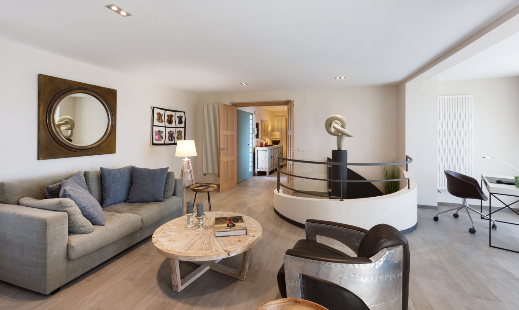 Salón moderno con sofá gris, sillón de estilo aviador y escalera de caracol con escultura central. Incluye zona de despacho y suelos de madera clara.