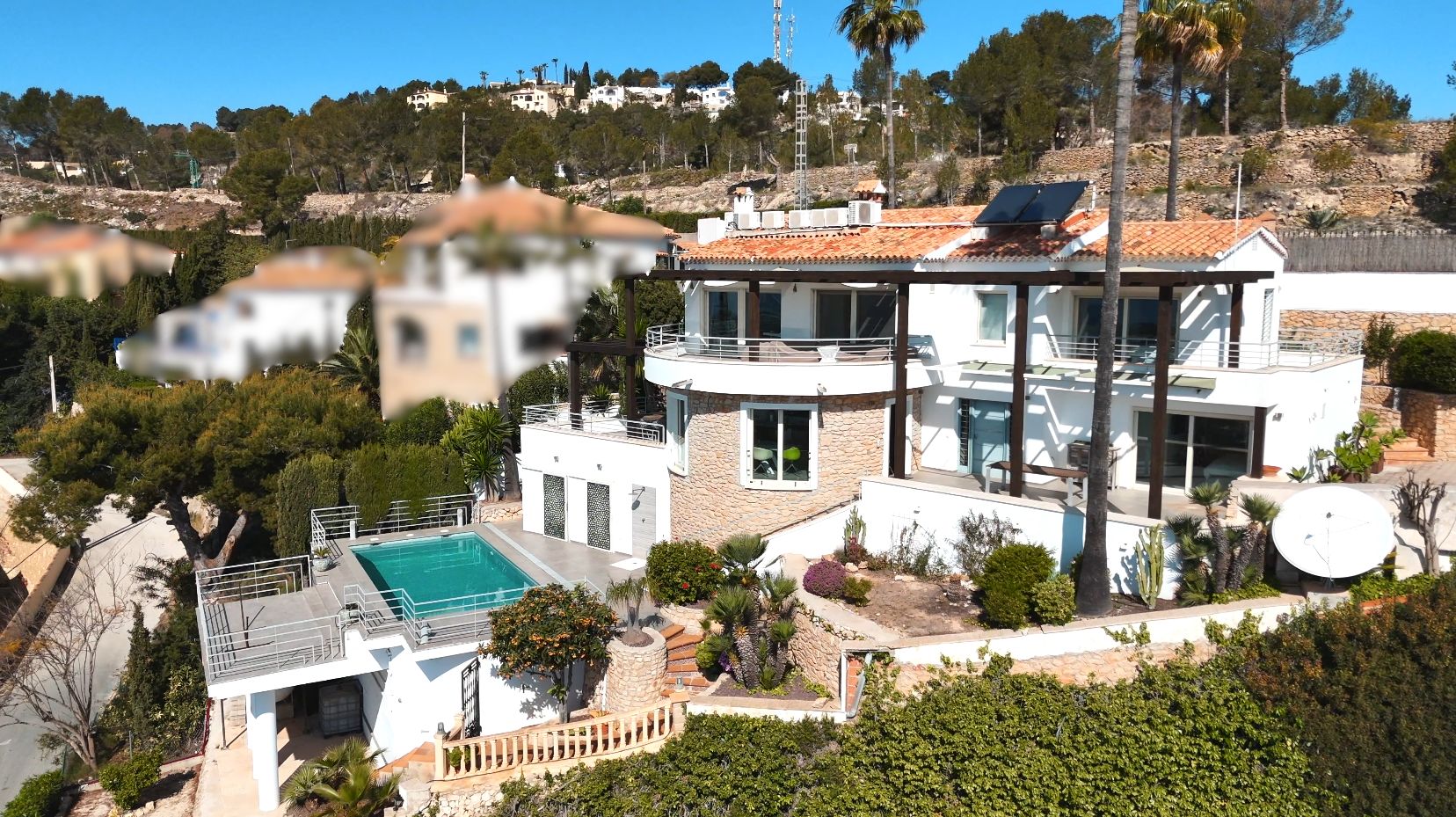 Villa mediterránea de varios niveles con fachada blanca, detalles en piedra, paneles solares y piscina privada rodeada de vegetación y palmeras.
