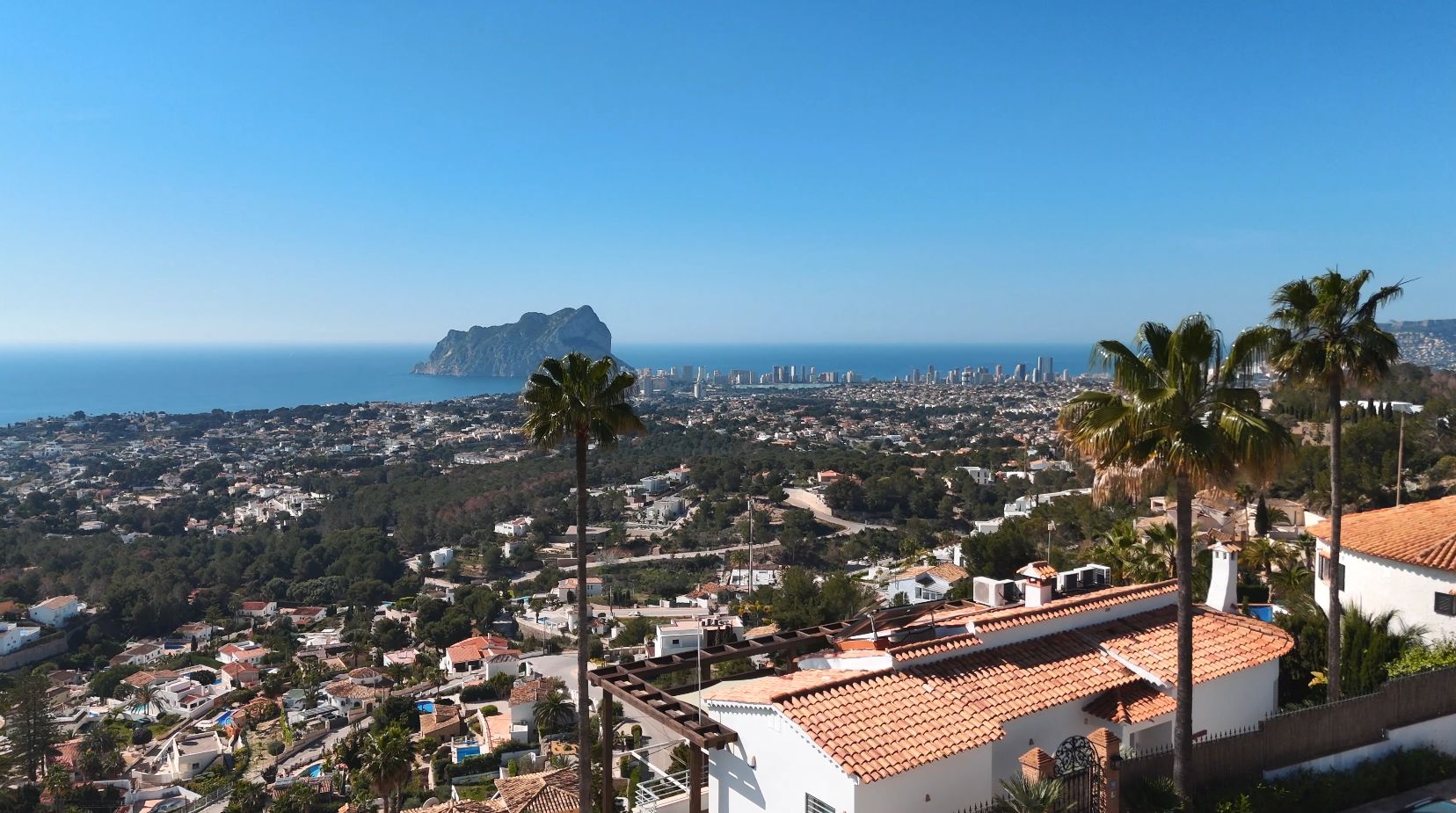 Vista panorámica elevada hacia el Peñón de Ifach y el skyline de Calpe, con villas mediterráneas de tejados cerámicos y palmeras en primer plano.