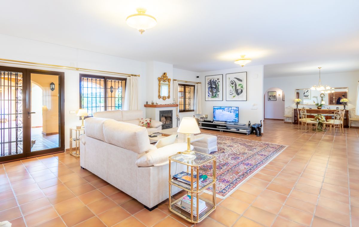 Amplio salón con suelos de terracota, sofá rinconero en tono crema, chimenea con espejo dorado y zona de comedor integrada con mesa de cristal.