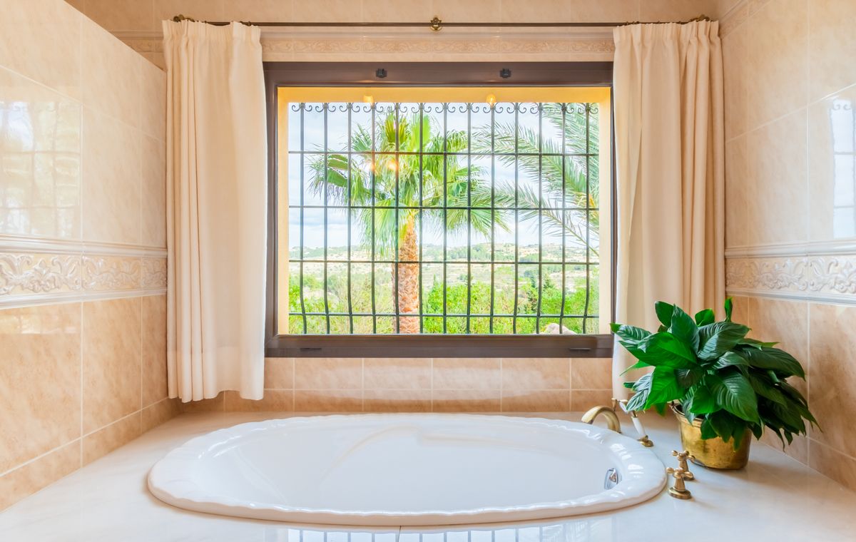 Bañera ovalada encastrada con grifería de latón, situada bajo un ventanal con rejas de forja y vistas despejadas a palmeras y al paisaje rural.