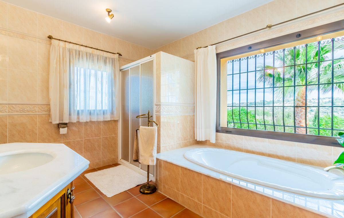 Baño con bañera integrada bajo ventana con rejas, cabina de ducha independiente, encimera de mármol blanco y suelos de terracota rústica.