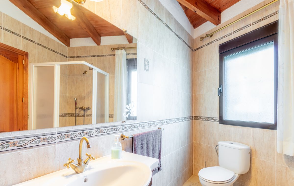 Baño con techo de madera inclinada y vigas vistas, paredes alicatadas en tonos beige y grifería de latón de estilo clásico.