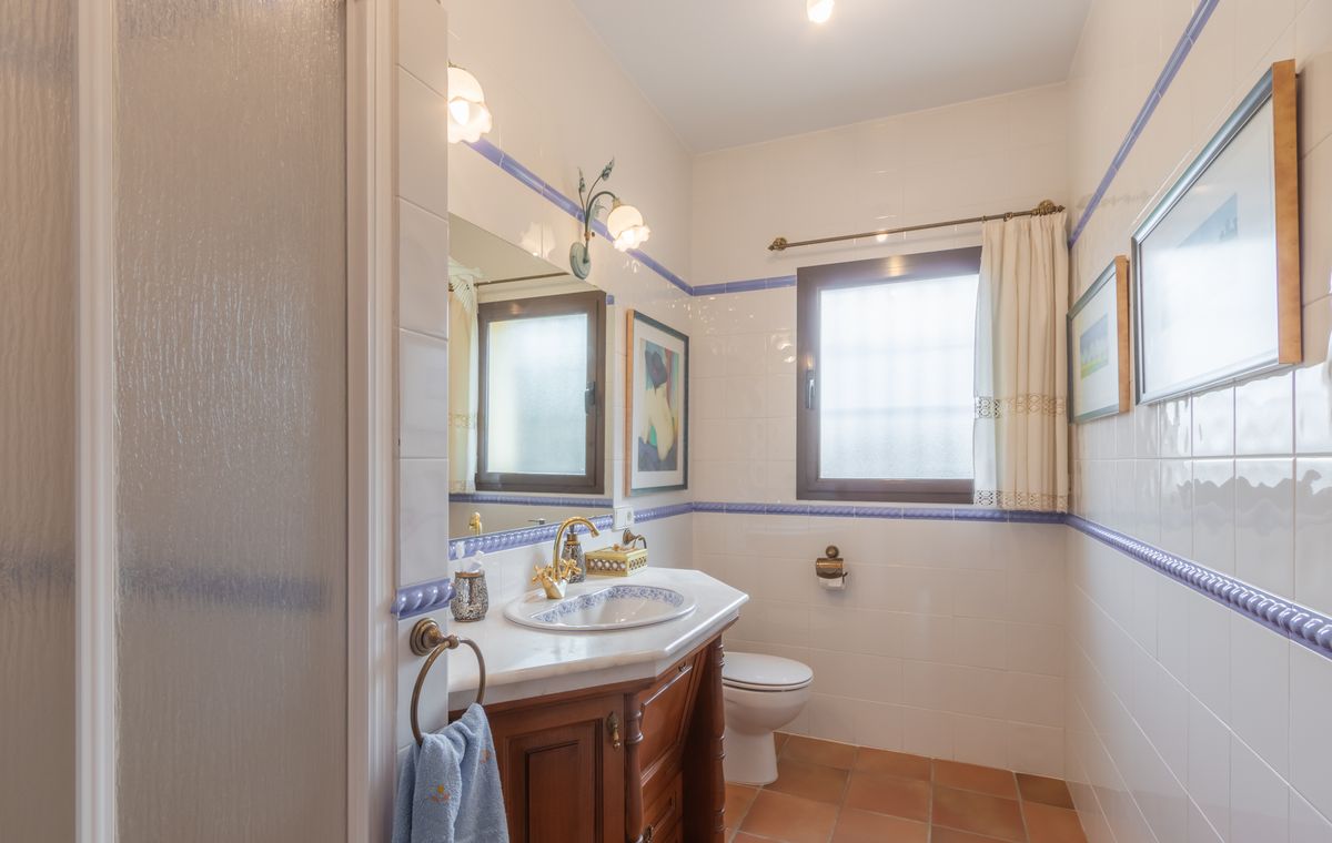 Baño de estilo tradicional con azulejos blancos, detalles en azul, mueble de madera tallada con encimera de mármol y suelos de barro cocido.