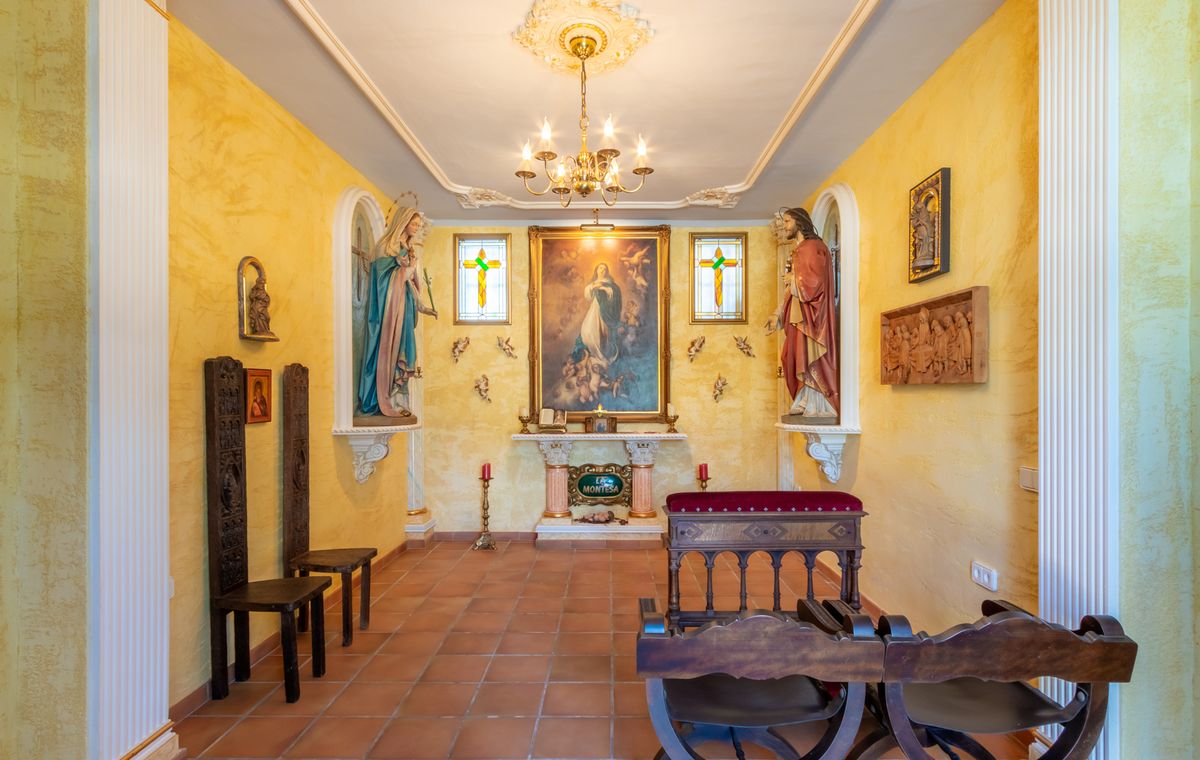 Capilla privada con paredes amarillas texturizadas, suelo de terracota, altar central con pinturas religiosas, hornacinas con estatuas y mobiliario de madera tallada.
