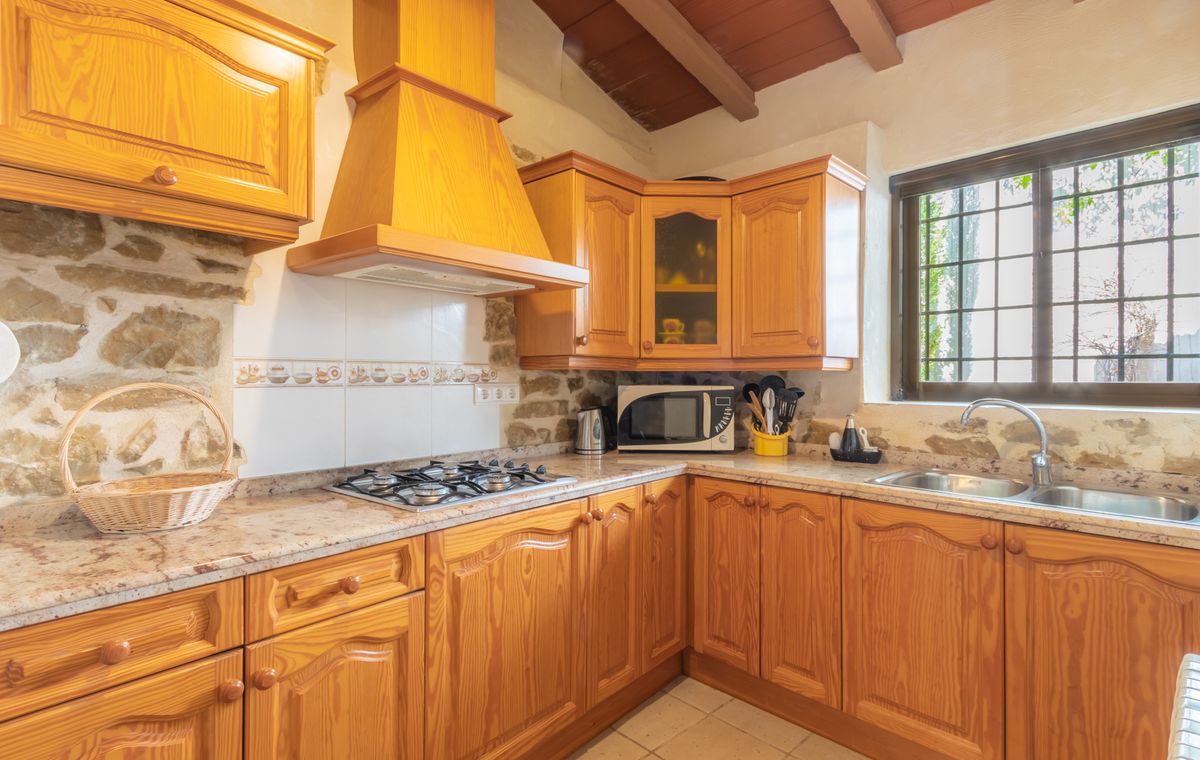 Cocina rústica con armarios de madera clara, paredes de piedra vista y encimera de granito. Destacan las vigas de madera en el techo y la campana extractora a juego.