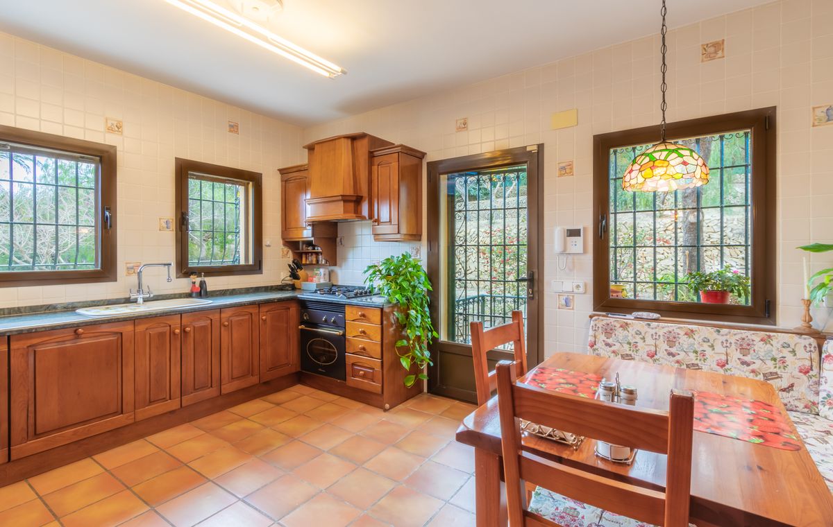 Cocina rústica con armarios de madera oscura, suelos de terracota y zona de comedor integrada con lámpara estilo Tiffany y bancos con motivos florales.