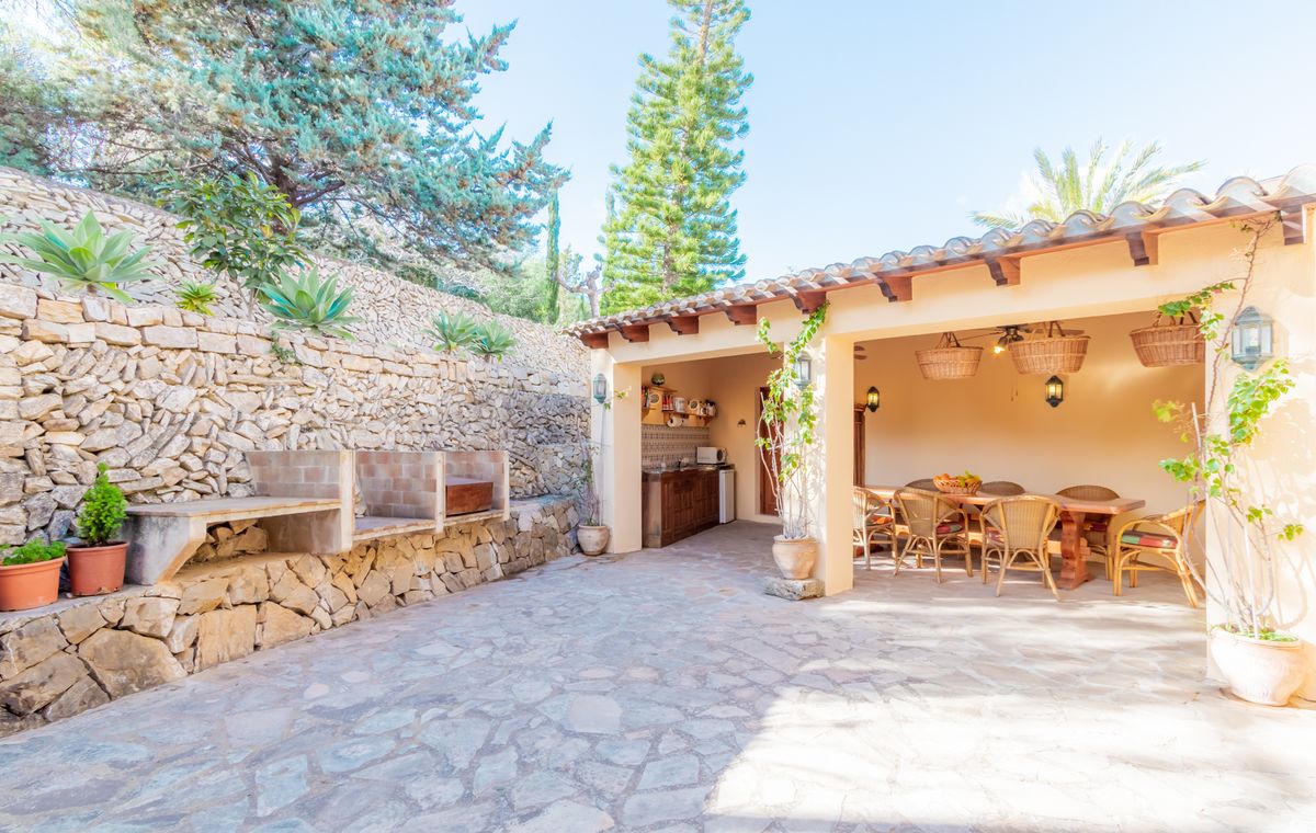 Comedor exterior con patio de piedra, barbacoa de obra y porche cubierto con vigas de madera, mobiliario de mimbre y una zona de cocina integrada.
