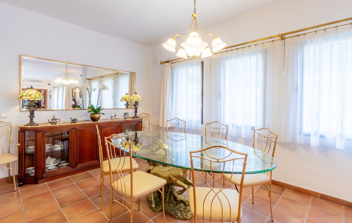 Comedor formal con mesa ovalada de cristal sobre base escultórica de latón, sillas doradas, suelo de terracota y un aparador de madera con espejo.