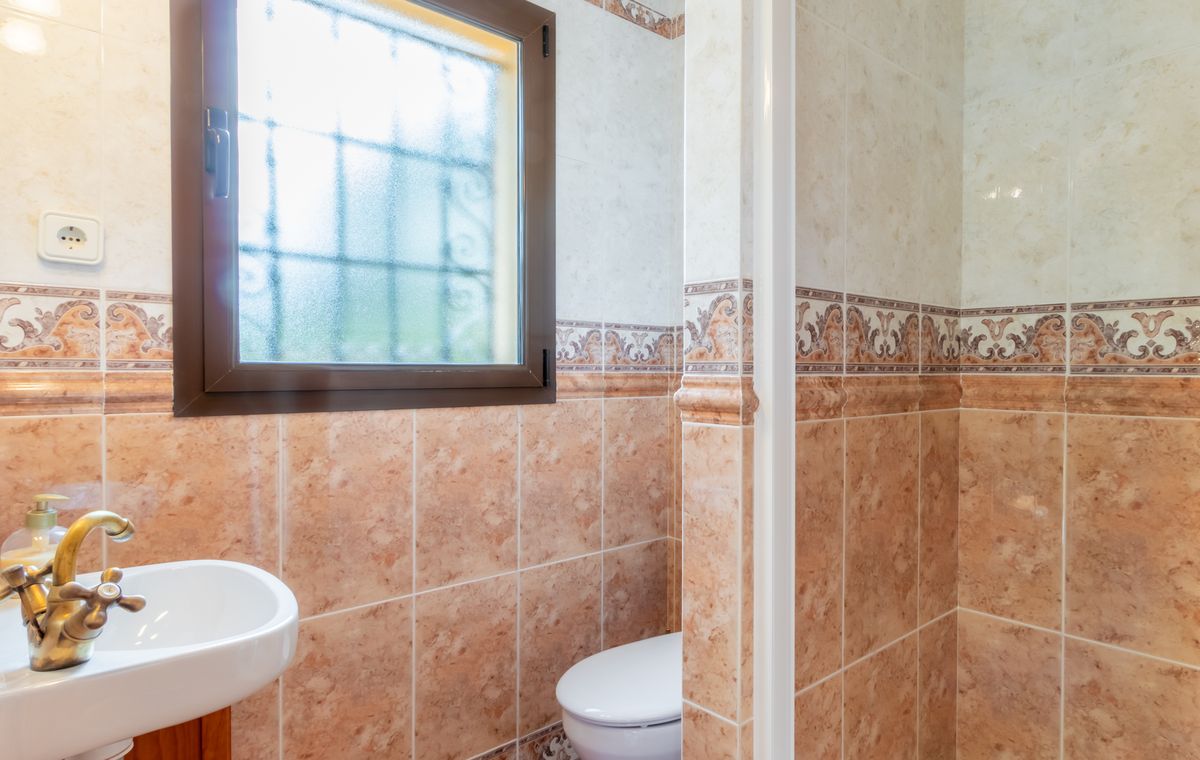 Cuarto de baño con azulejos de efecto mármol en tonos tierra, grifería de latón de estilo clásico y ventana de cristal translúcido con reja de seguridad.